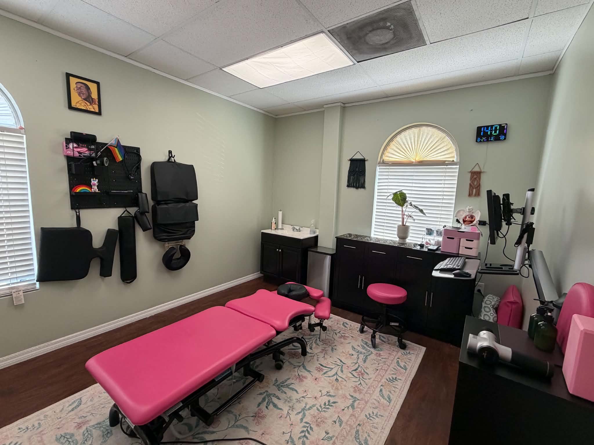 A room with pink examination tables, a patterned rug, a small black cabinet, a plant, a wall clock showing 2:40, and various decorations on the wall, including a framed portrait and wall hangings. There are windows with blinds on two sides of the roo