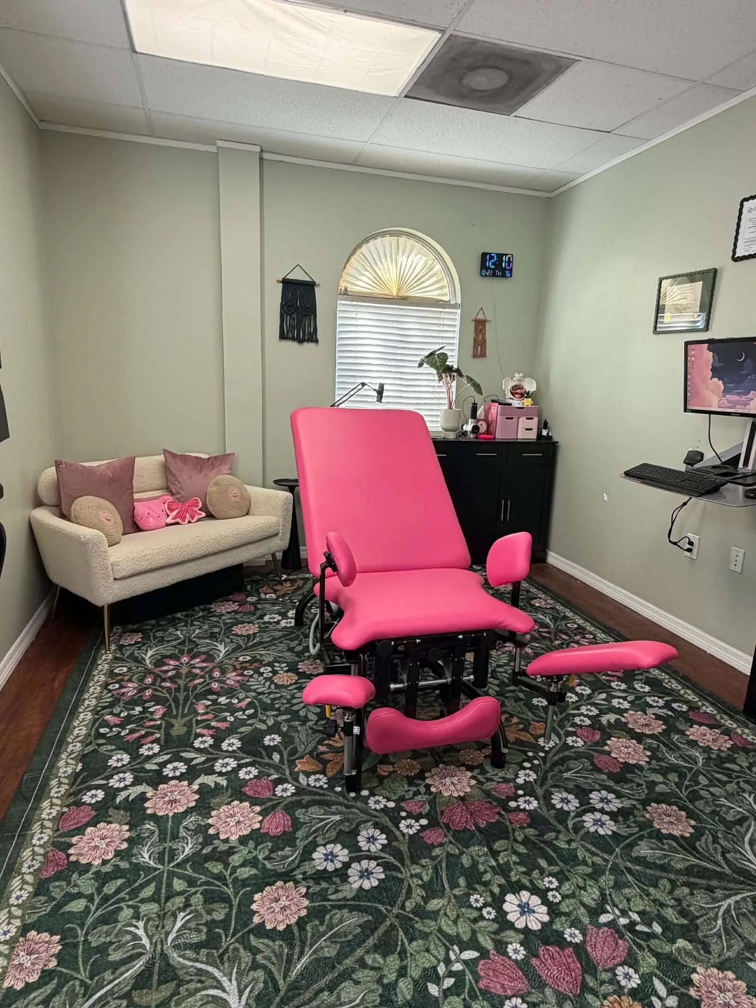 A medical or chiropractic examination room with a pink treatment table, pink stool, and a white and wood rolling cart with blankets and supplies. There is a black bookcase filled with books and decorative items, a printer on the corner of a dark wood cabinet, a computer with a pink and purple moon and clouds wallpaper on the monitor, and a potted plant on the cabinet. The room has a light-colored wall, dark wood flooring, and a floral rug under the treatment table.