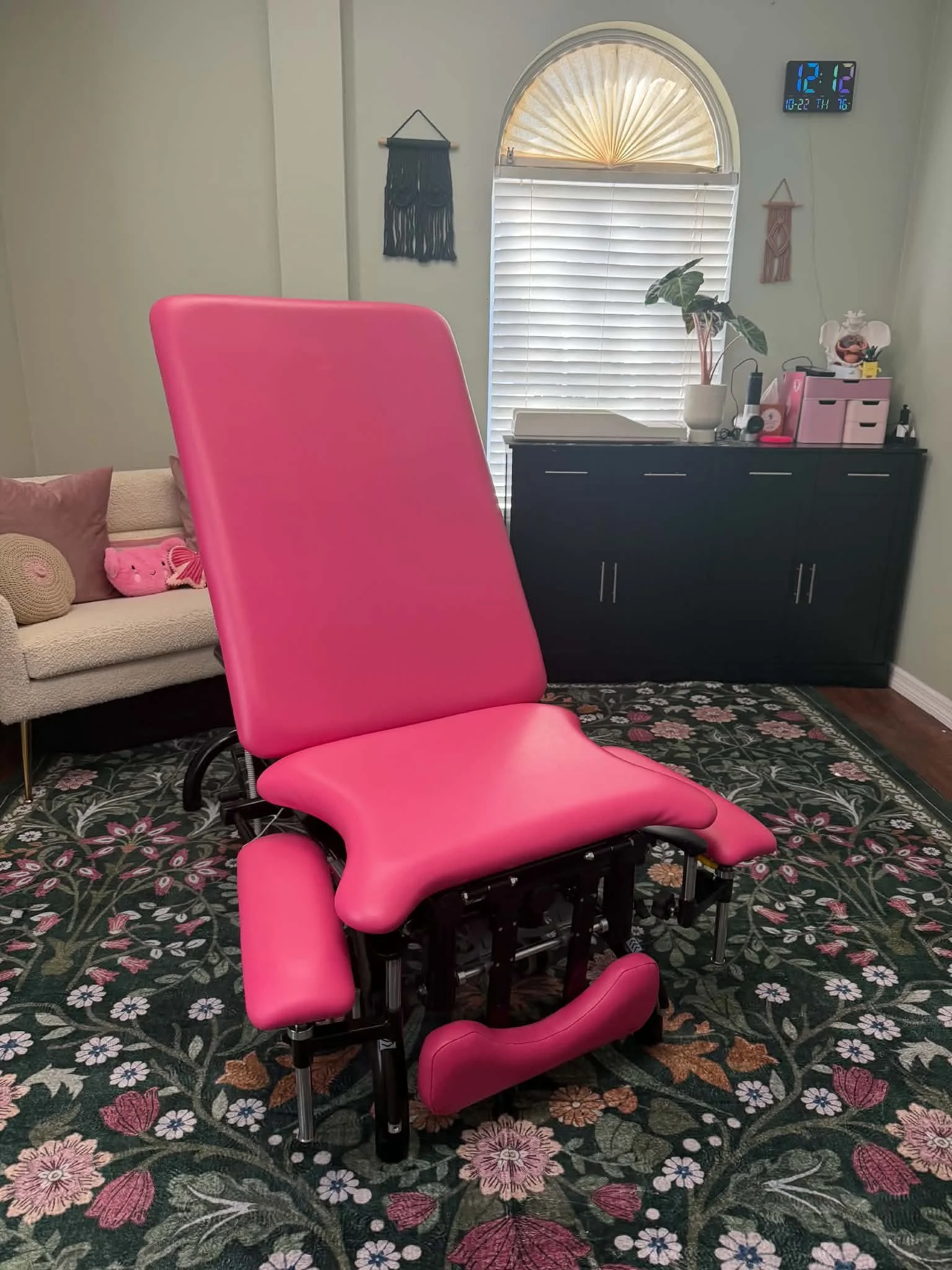 A pink medical examination chair in a clinic room, with a white sofa and decorative pillows, a small table with medical supplies, and wall decorations including a framed butterfly picture and the word 'Lymos' on the wall.