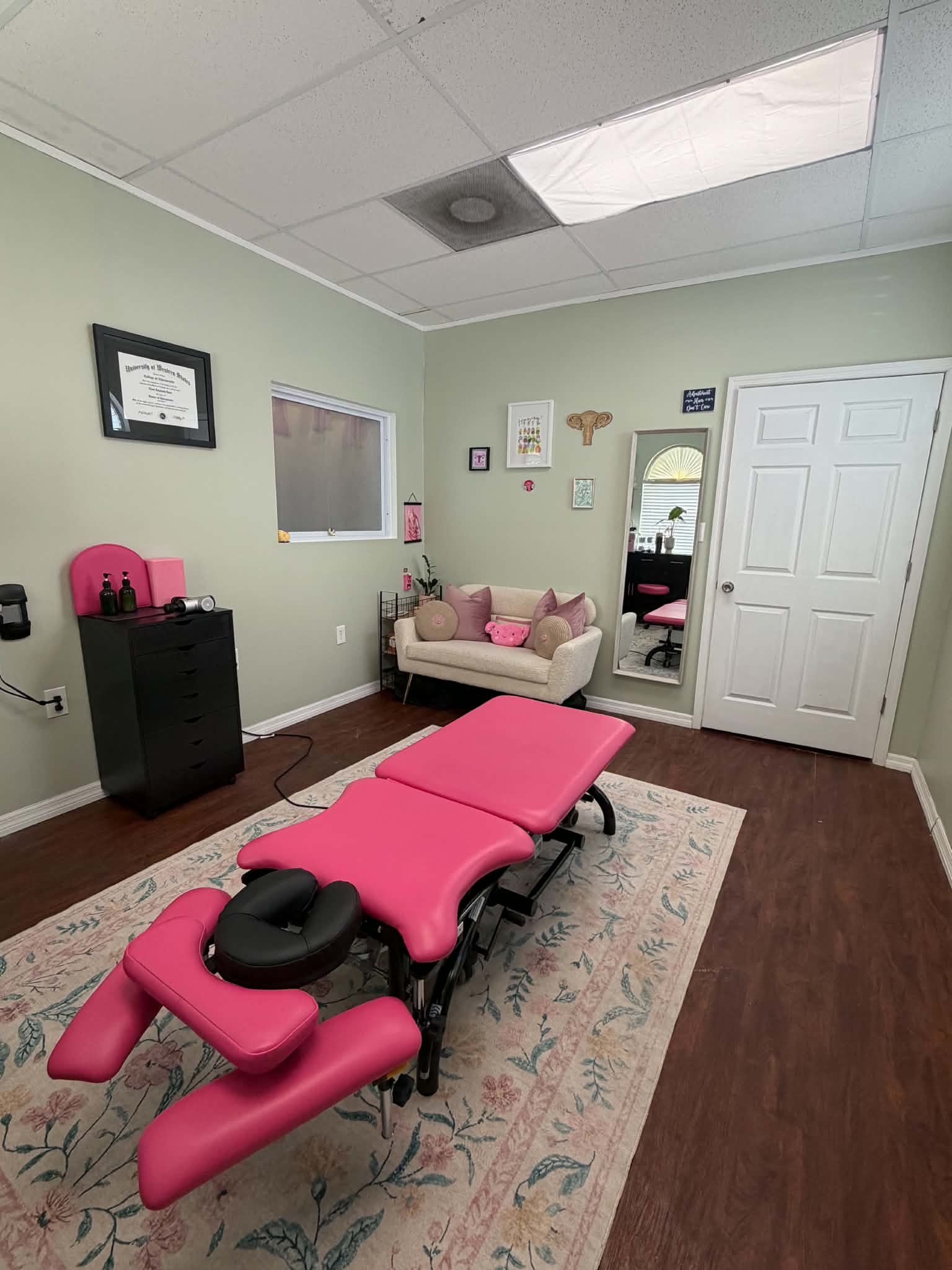 A pink massage or chiropractic table with black head support in a room with light green walls, hardwood floors, a patterned area rug, a small sofa with pink pillows, wall decorations, a mirror, and medical certificates.