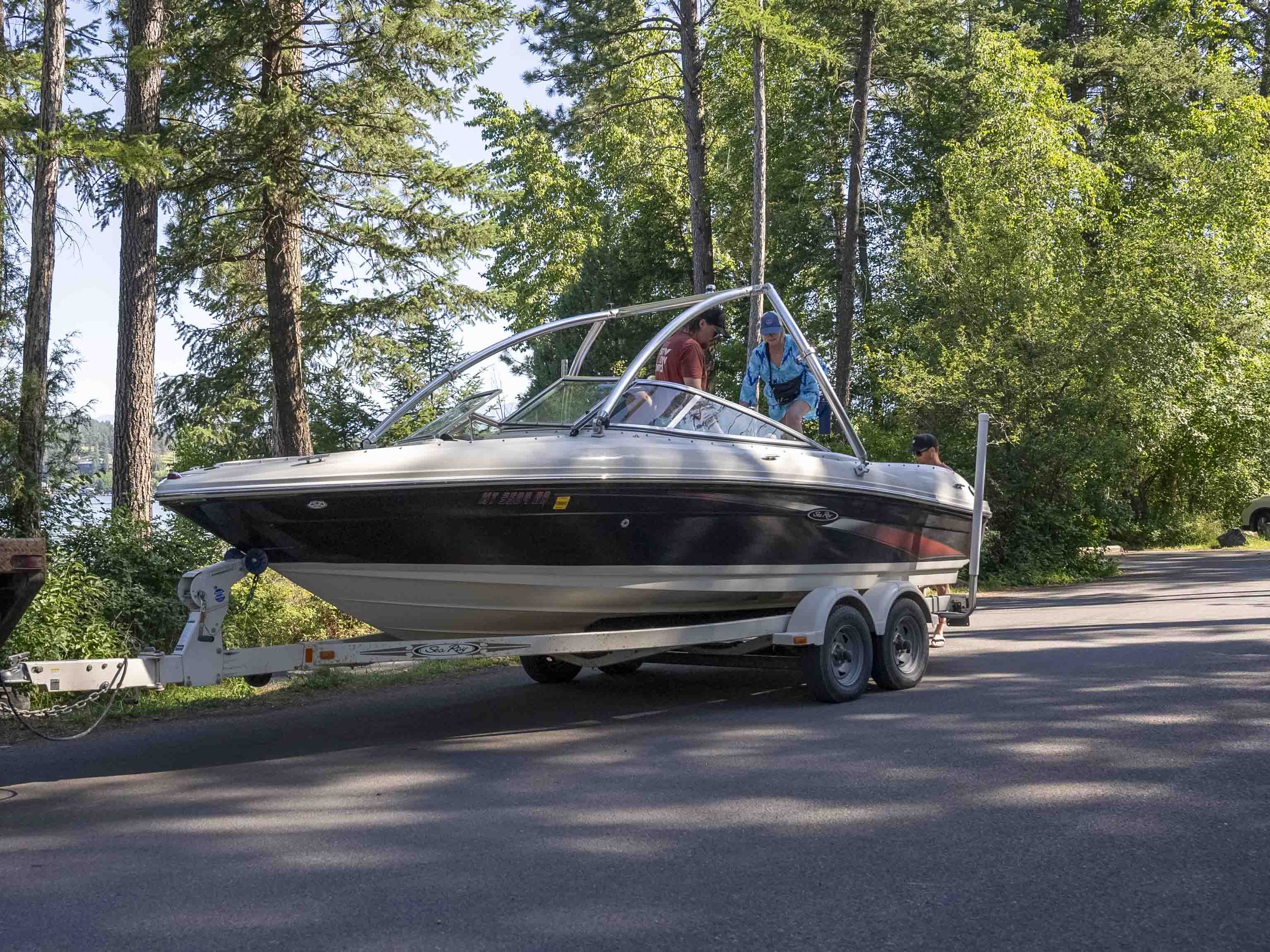A group of people in casual attire standing on a boat on a trailer, preparing for a boat ride on a road near a wooded area with trees and a glimpse of water in the background.