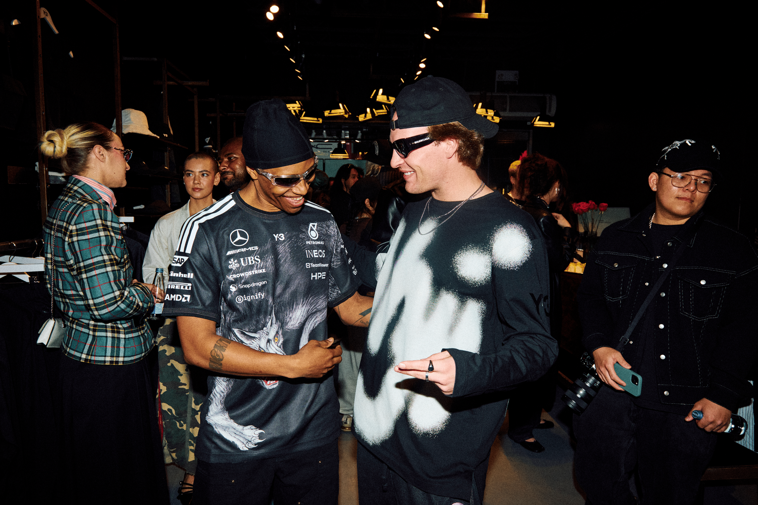 Two male campaign tastemakers dressed in Adidas Y-3 in the center of the image are smiling and chatting; both men are wearing black and white shirts from Y-3's Spring/Summer 2026 collection and dark sunglasses; in a lively social gathering.