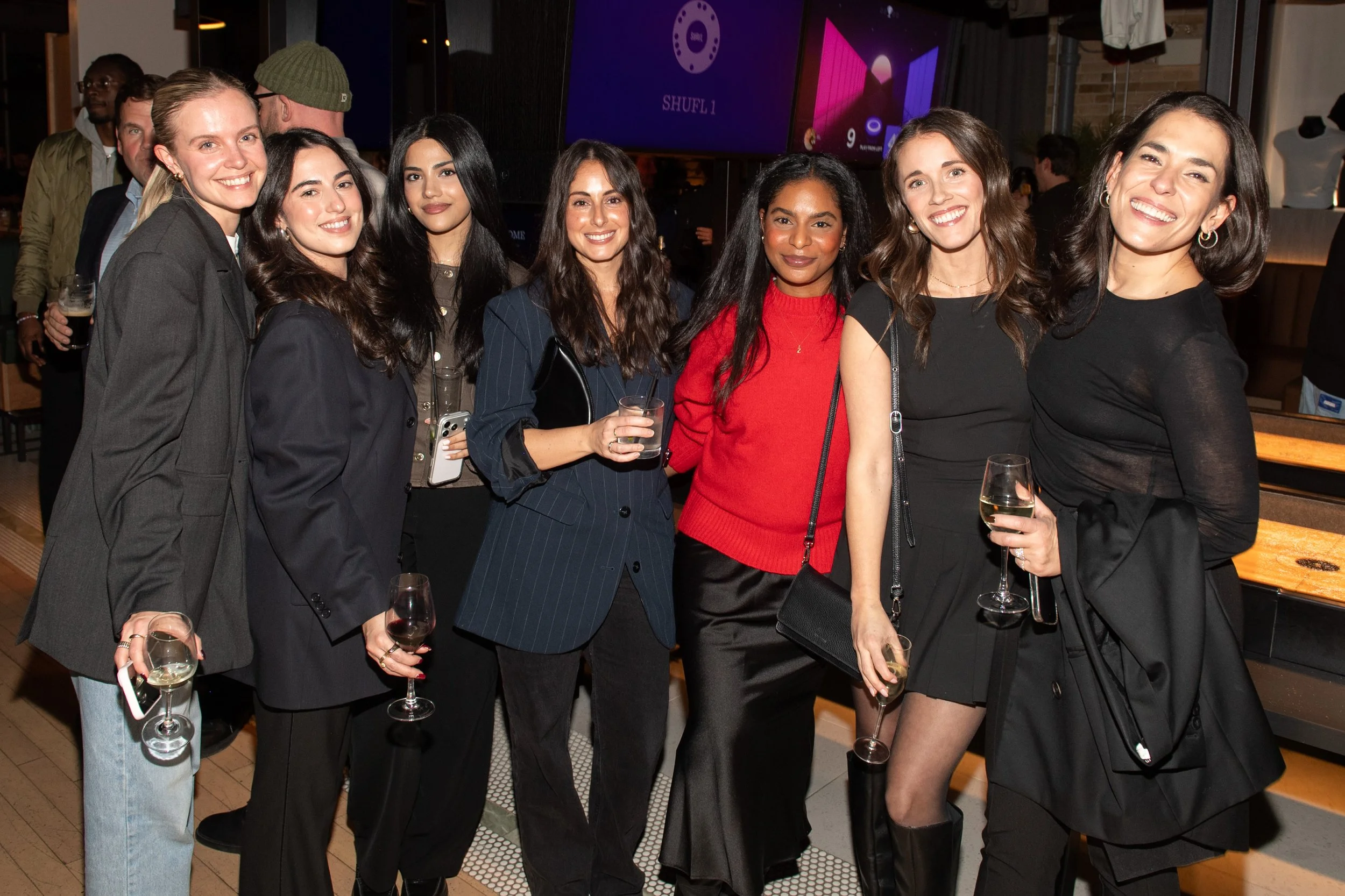 A group photo of 7 women at the MNTD launch event hosted by Public/Realm; six women are wearing black, while one woman stands out in a red blouse and black silk skirt