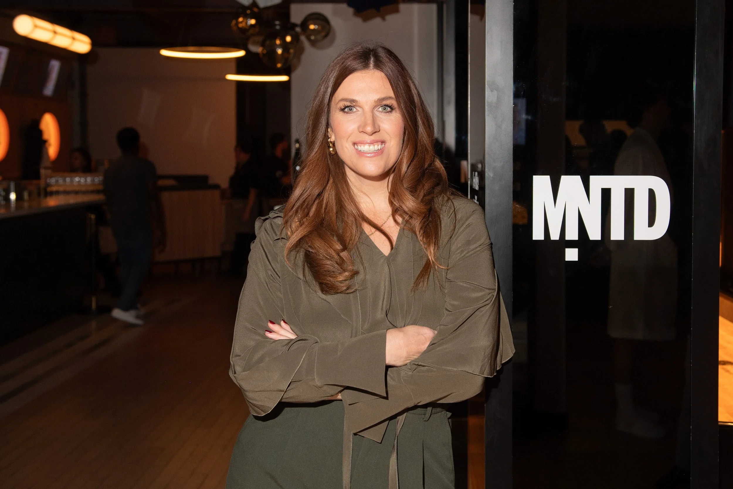 Knix Founder & President, Joanna Griffiths at the launch event for MNTD Menswear; Joanna is standing with her arms crossed, smile on her face, next to a MNTD sign