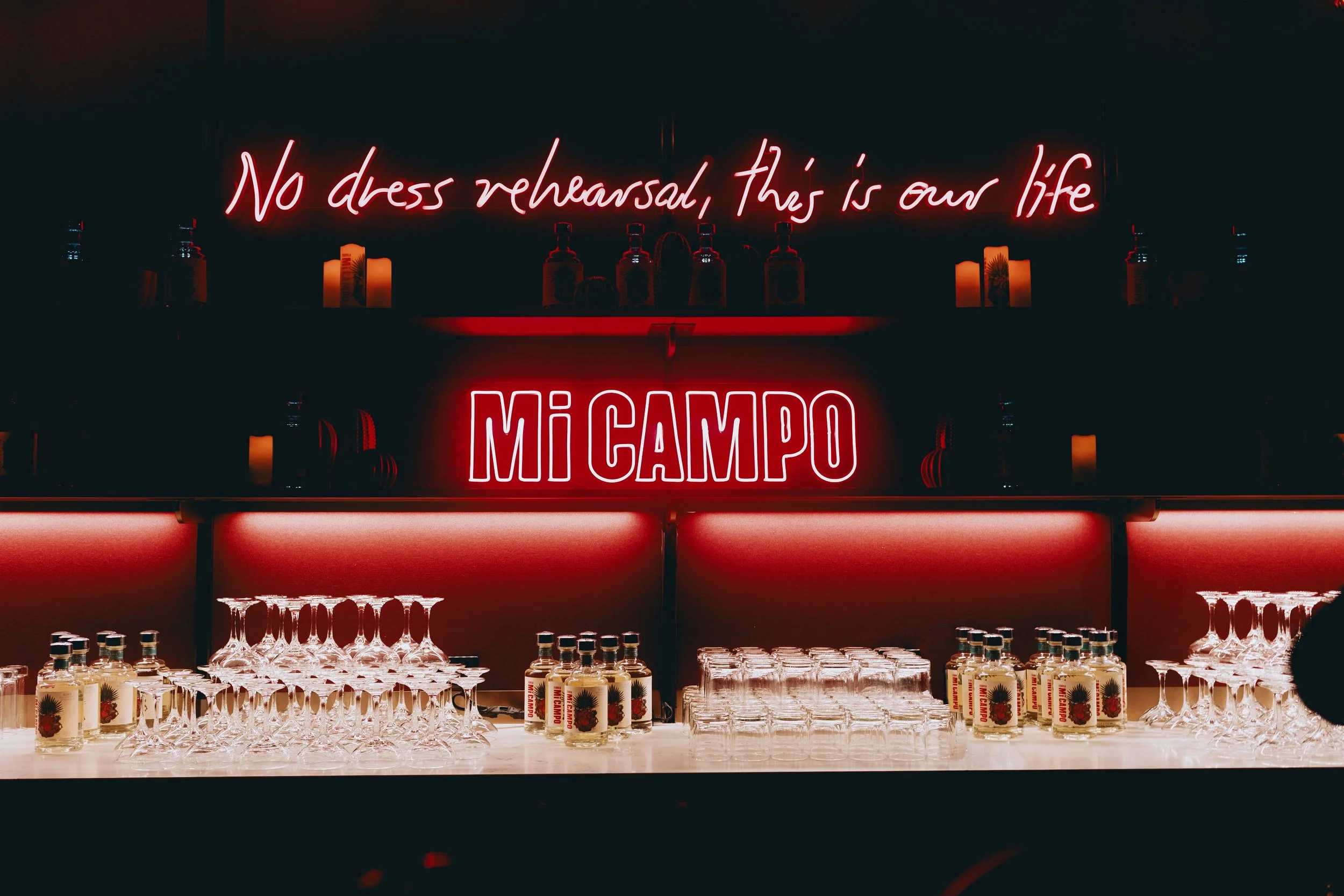 A wide photo of a glowing backbar with a neon sign that reads "no dress rehearsal, this is our life", and underneath it is another neon sign that says Mi Campo; Public/Realm created this immersive brand experience for Mi Campo tequila