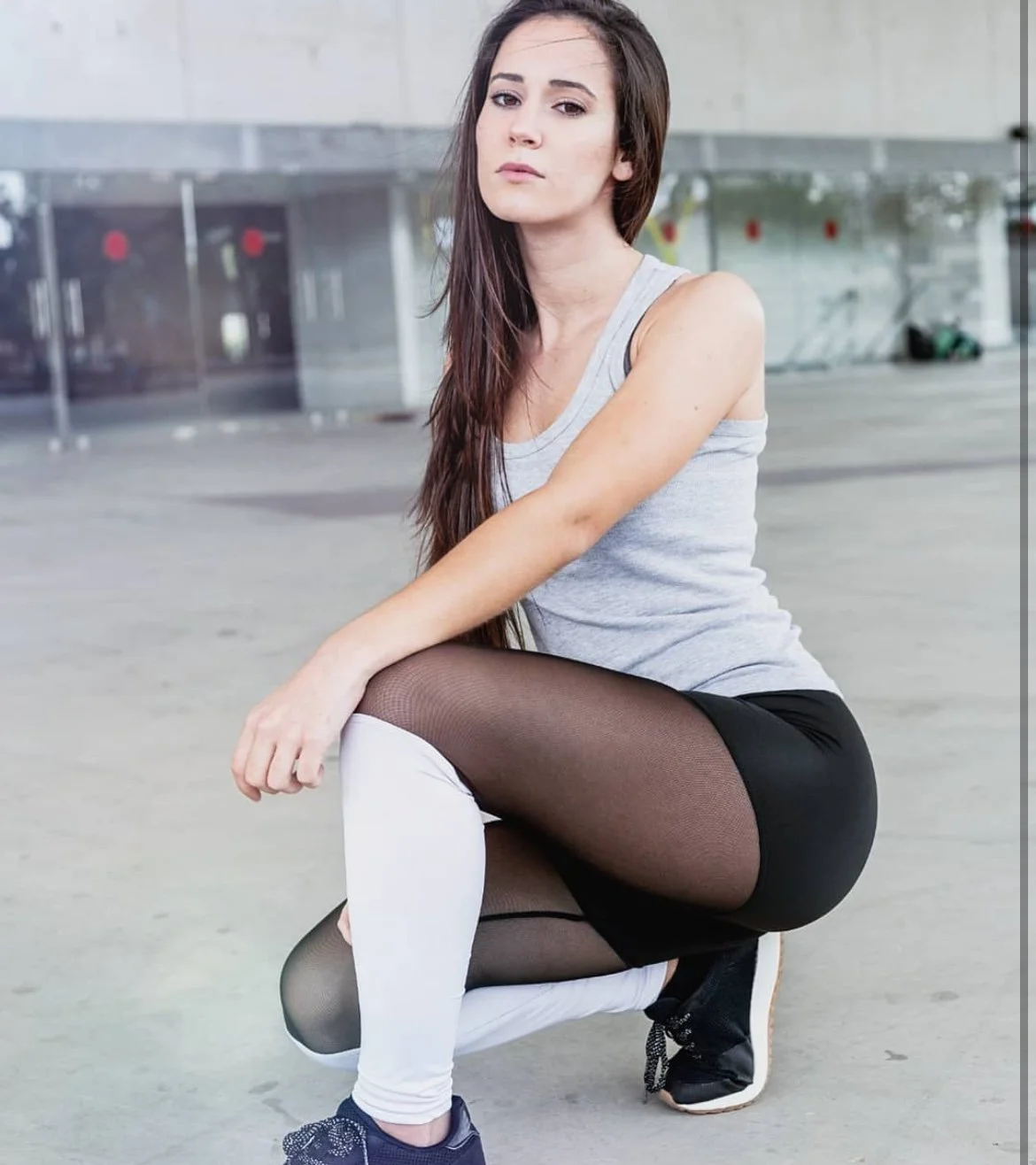 Joven mujer en ropa deportiva, en posición de cuclillas, en un espacio abierto, con fondo de un edificio modernista.