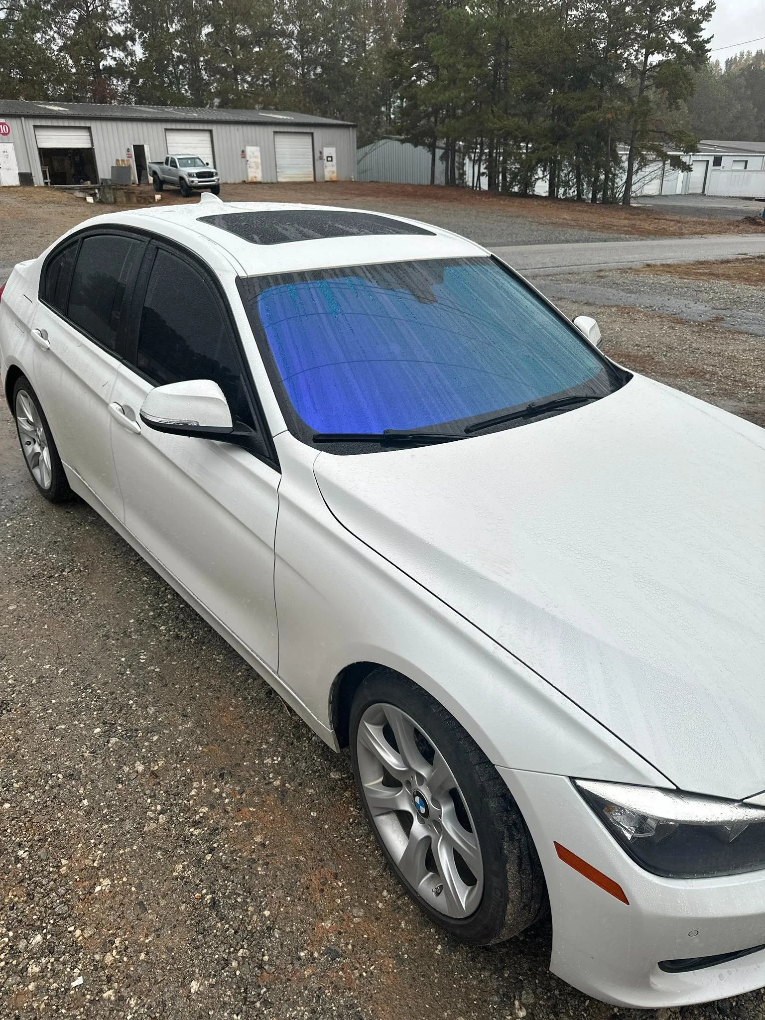 White BMW sedan parked on gravel driveway next to industrial buildings, rainy weather, overcast sky, trees in the background.