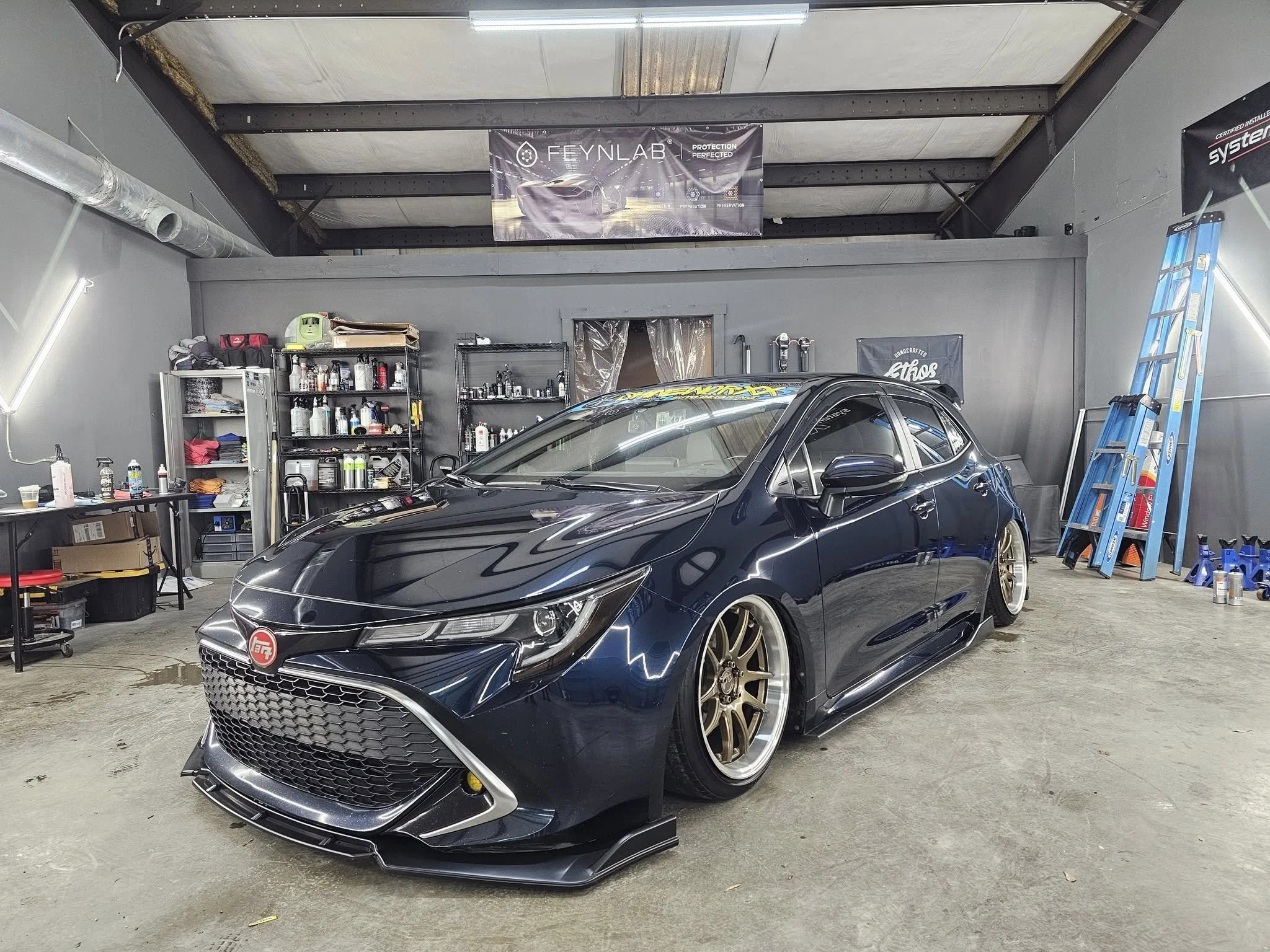 A dark-colored sports car with golden wheels inside a garage, surrounded by various automotive tools and supplies.