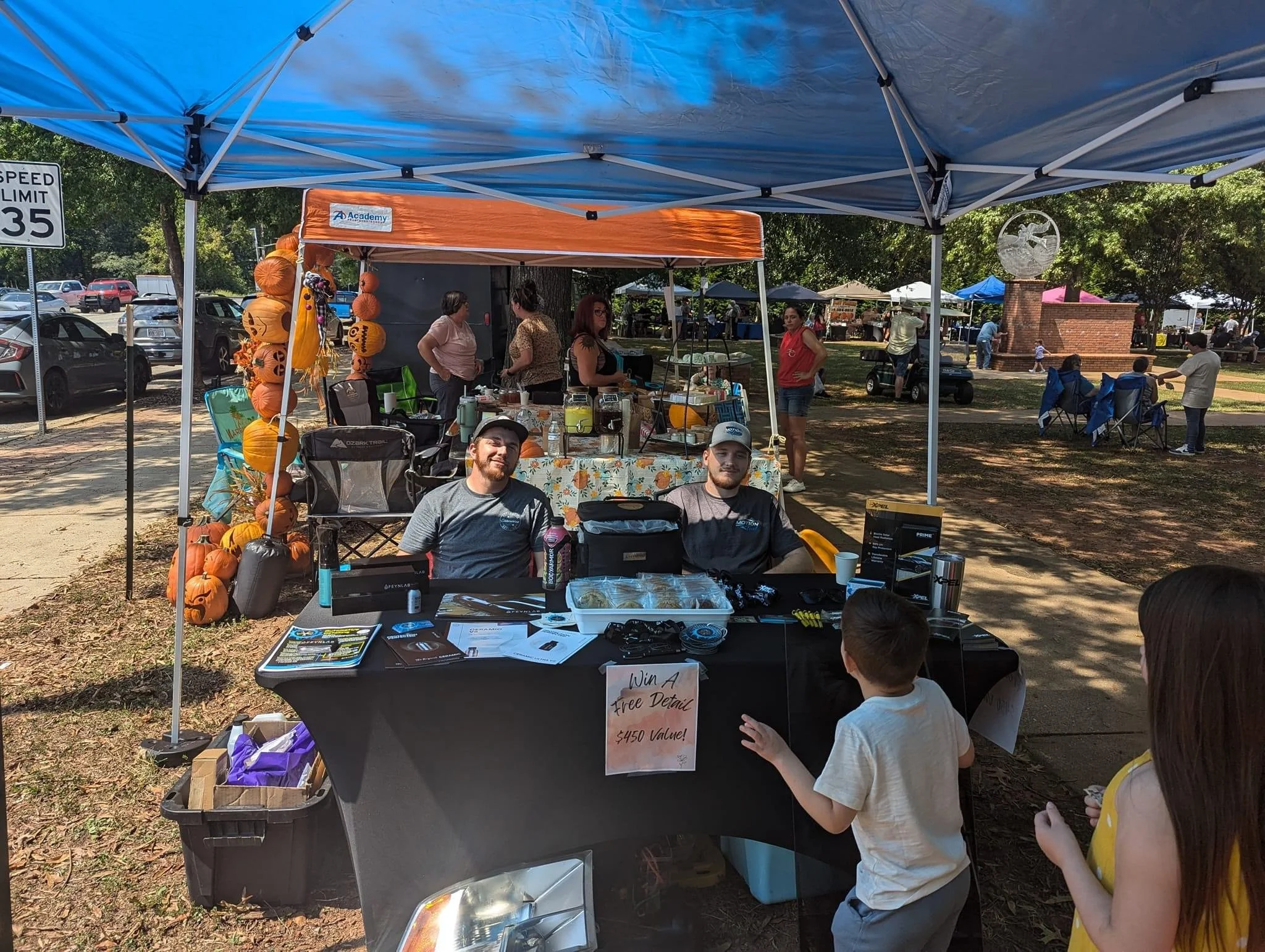 An outdoor market stall at a festival or fair with two young men sitting behind a table under a blue canopy, surrounded by children and adults. The table displays promotional materials, and there is a sign offering a free detail valued at $450. Behind the stall, other vendors and people are visible, along with trees and a park area.