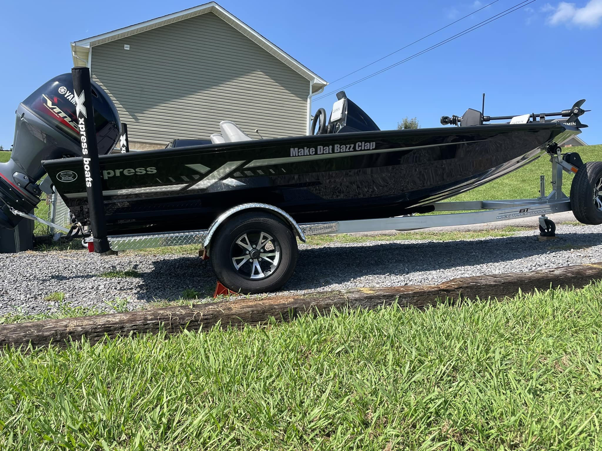 Black fishing boat on a trailer with a Yamaha outboard motor, parked on a gravel driveway beside a grassy yard with a house in the background.