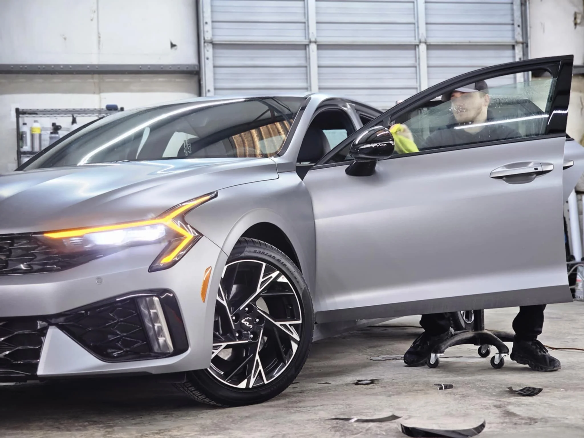 A man working on a silver luxury car inside a garage, sitting on a rolling stool, with the driver's side door open.