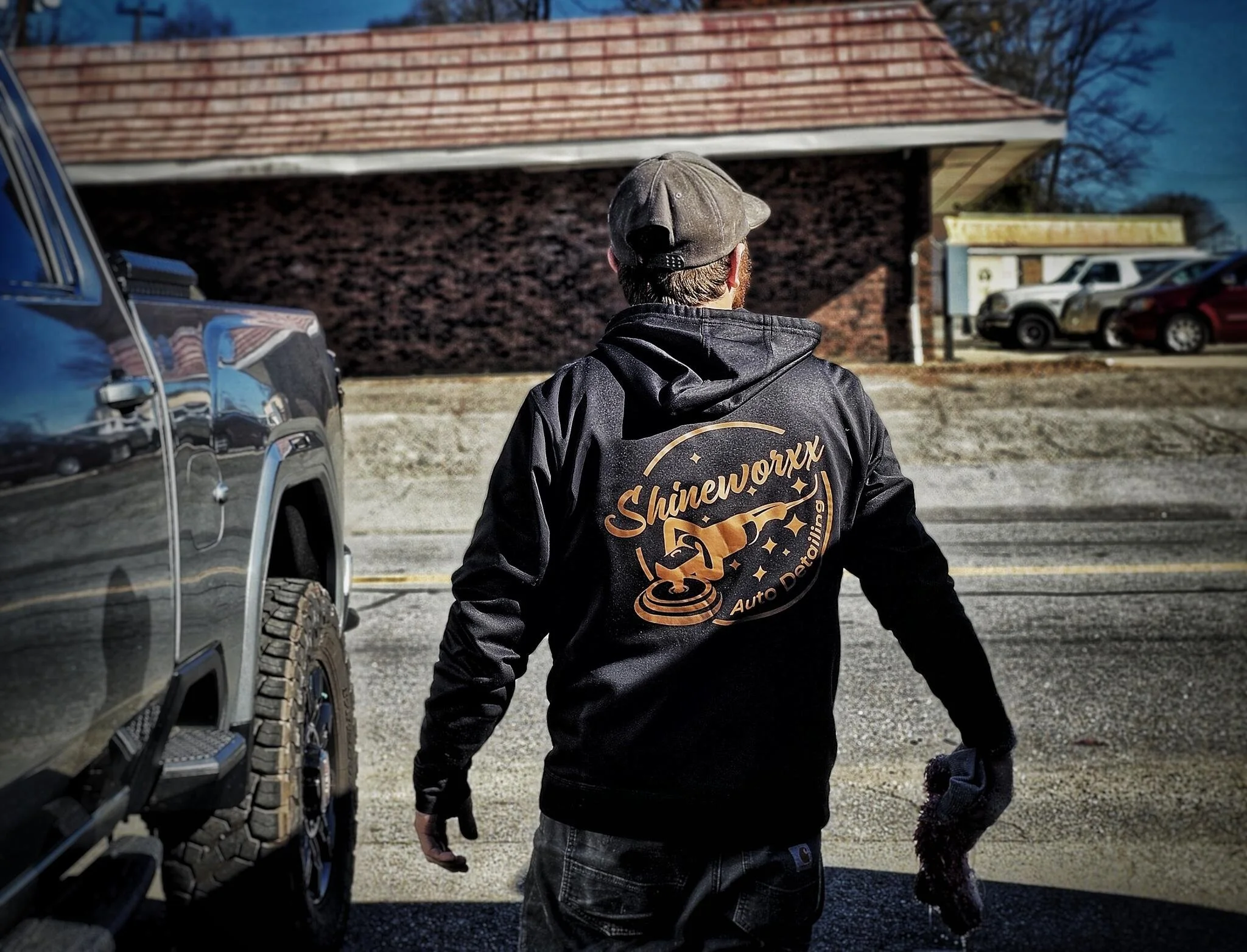 A person wearing a black jacket with 'Shineworkx Auto Detailing' logo on the back, gloves, and a cap, standing outdoors next to a parked vehicle on a street.