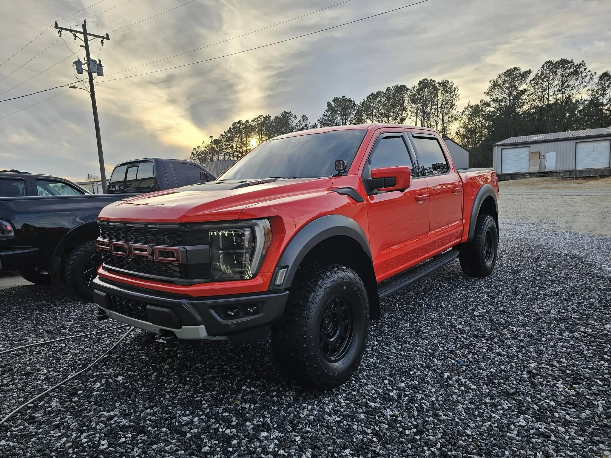 Red Ford truck parked on gravel lot with other vehicles and a building with white garage doors in the background, during sunset.