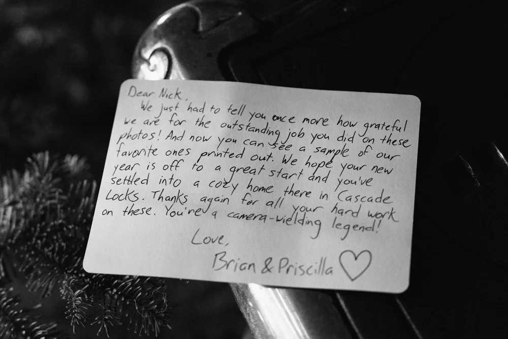 A handwritten thank-you note on a piece of paper, placed on a dark surface with a pine branch nearby. The note expresses gratitude for photography work and includes a heart drawing.