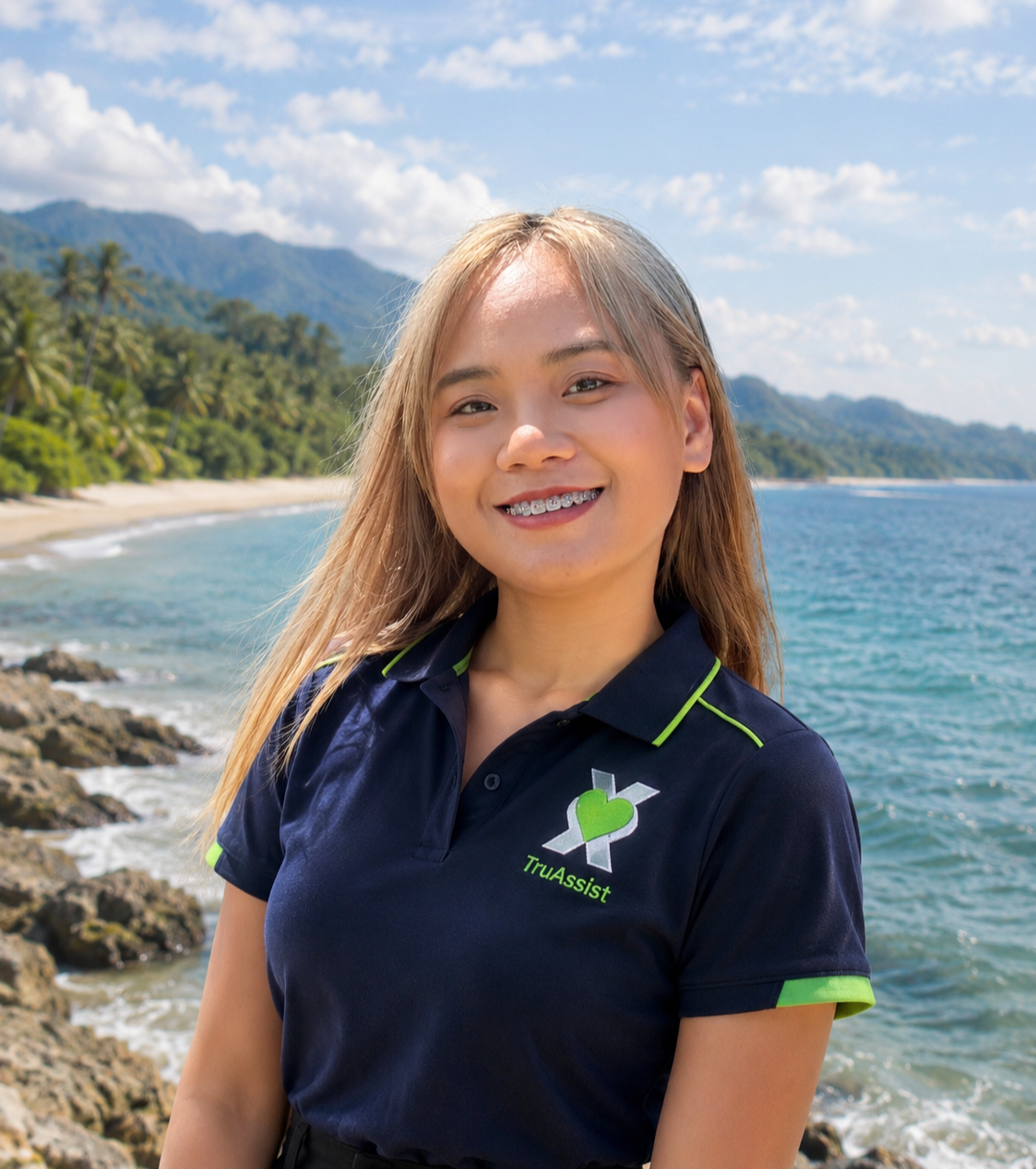 A young woman with blonde hair smiling at the camera, wearing a navy blue shirt with green accents and 'TruAssist' logo, standing near the beach with rocks, trees, and mountains in the background under a partly cloudy sky.