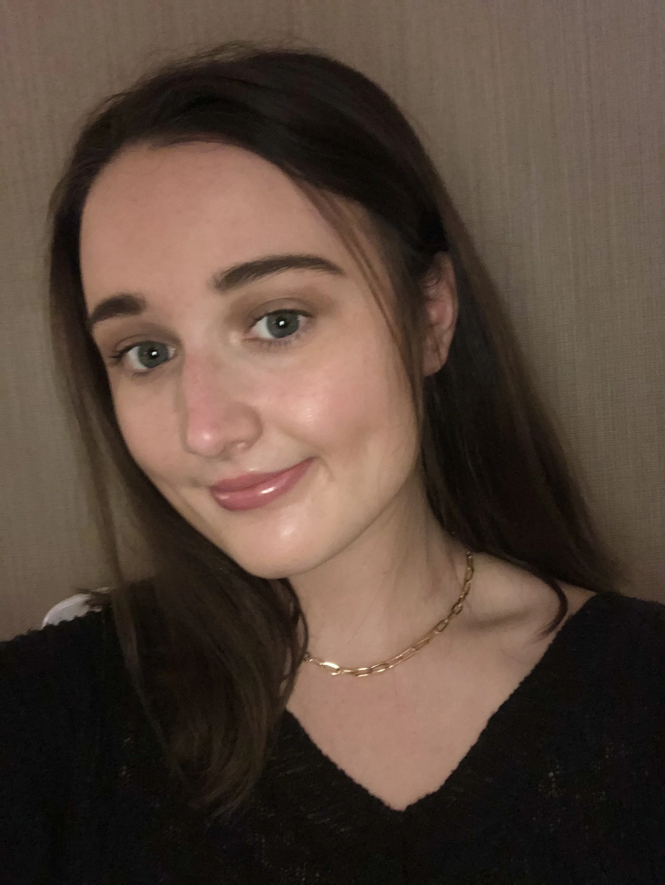 A young woman with long brown hair, fair skin, and blue eyes taking a selfie, wearing a black top and a gold chain necklace, standing against a neutral-colored wall.