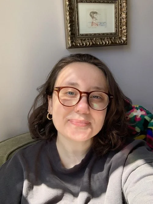 A woman with shoulder-length brown hair, wearing glasses and a black and gray sweatshirt, sitting on a couch with a blanket behind her, and a framed picture of a dog on the wall above.