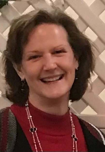 A woman with short brown hair smiling, wearing a red top, earrings, and a beaded necklace, standing in front of a white lattice background.