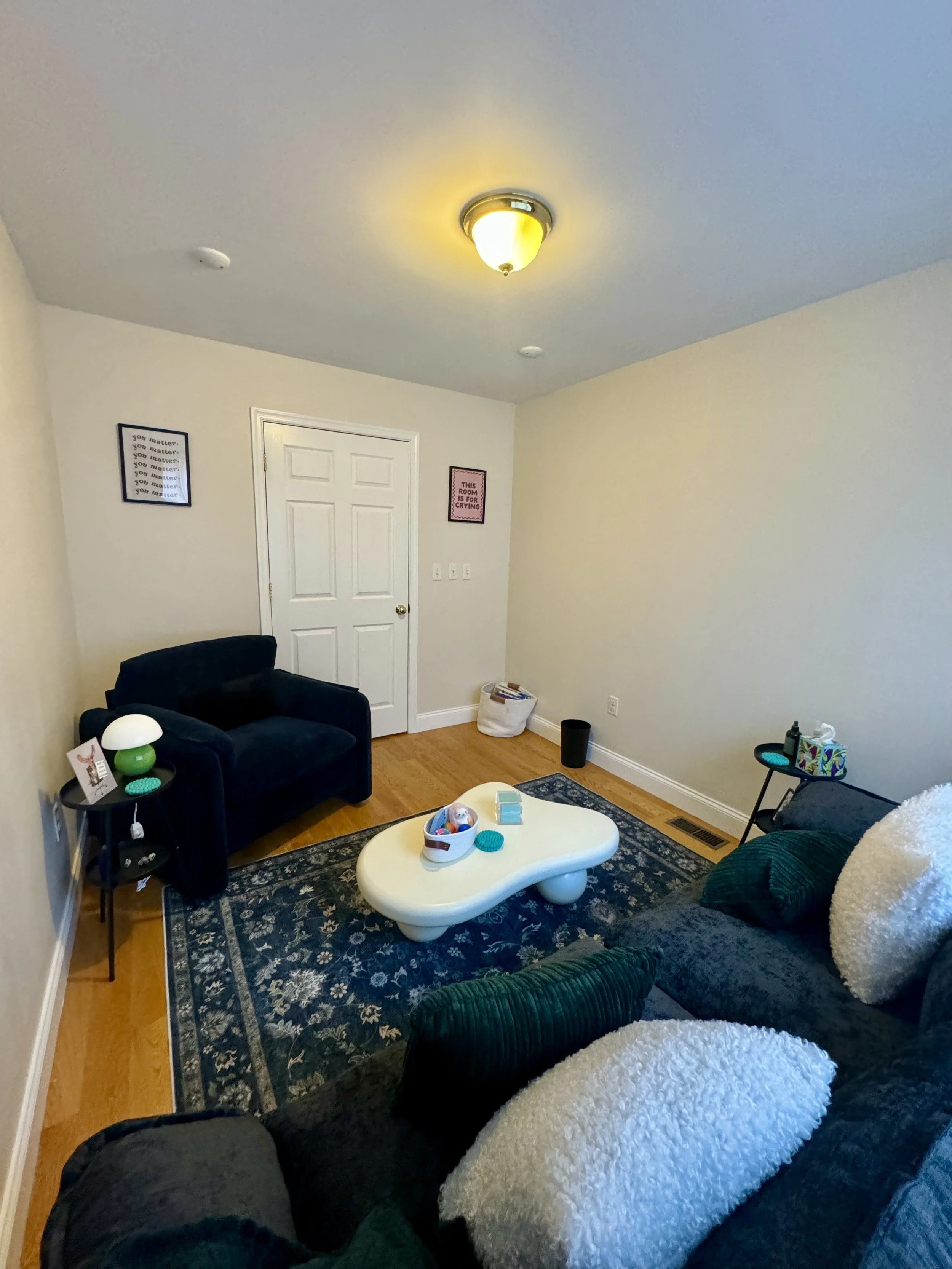 Living room with white walls, hardwood floors, a ceiling light, a white coffee table, a black armchair, a dark gray sofa with white pillows, two framed posters on the wall, a small side table with a lamp, and a door at the back.