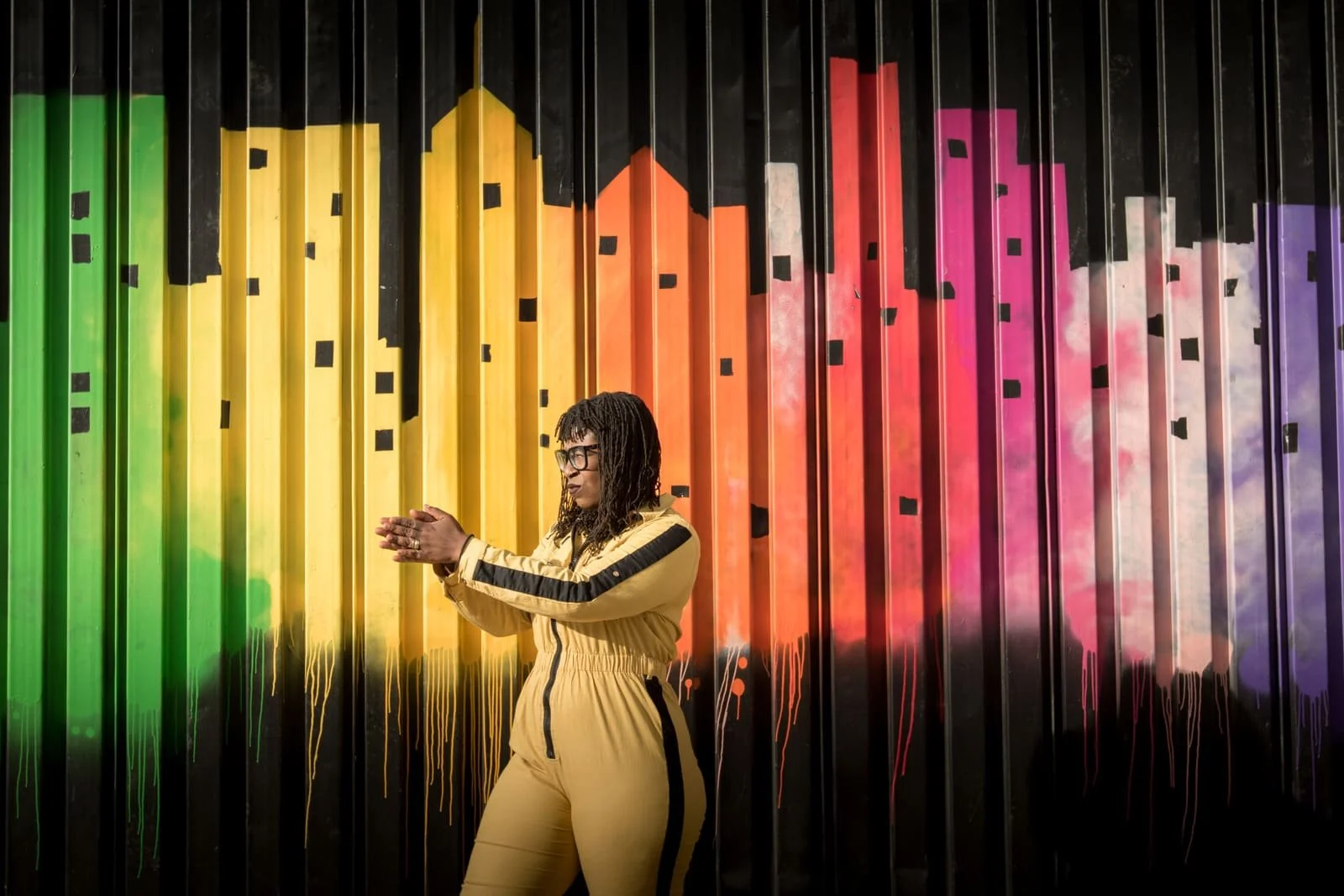 Person in yellow jacket and pants clapping in front of a black wall with colorful rainbow and cloud graffiti