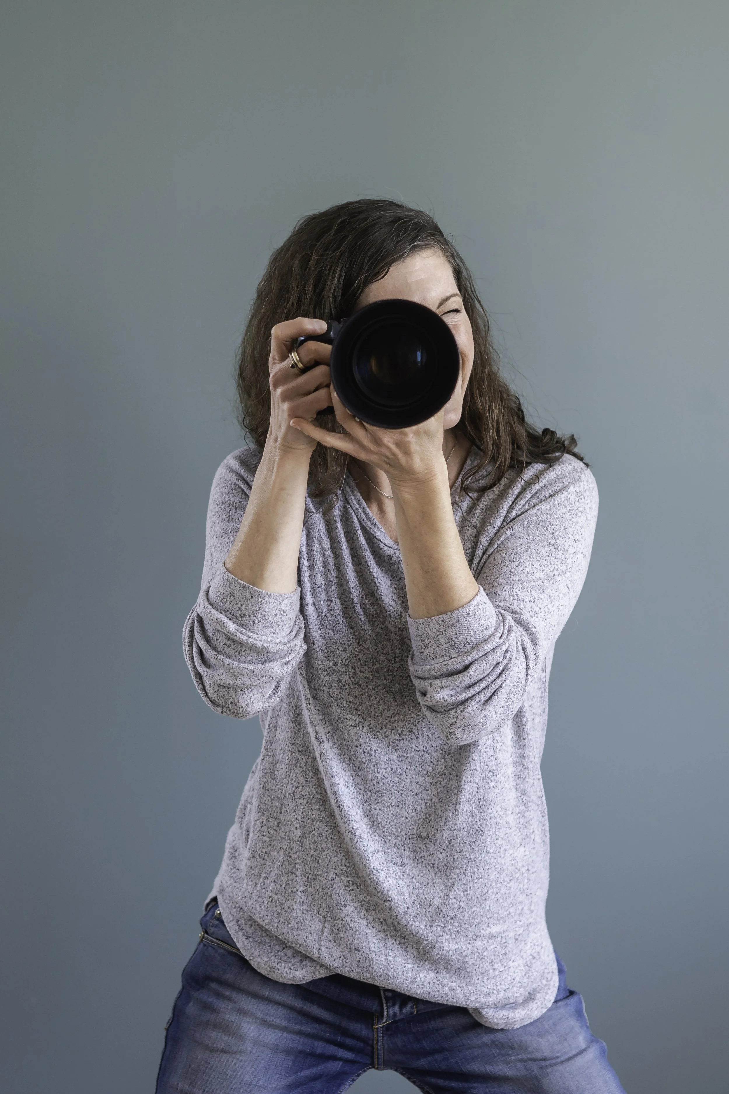 A woman with wavy brown hair, wearing a gray long-sleeve shirt and blue jeans, taking a photo with a DSLR camera against a plain green backdrop.
