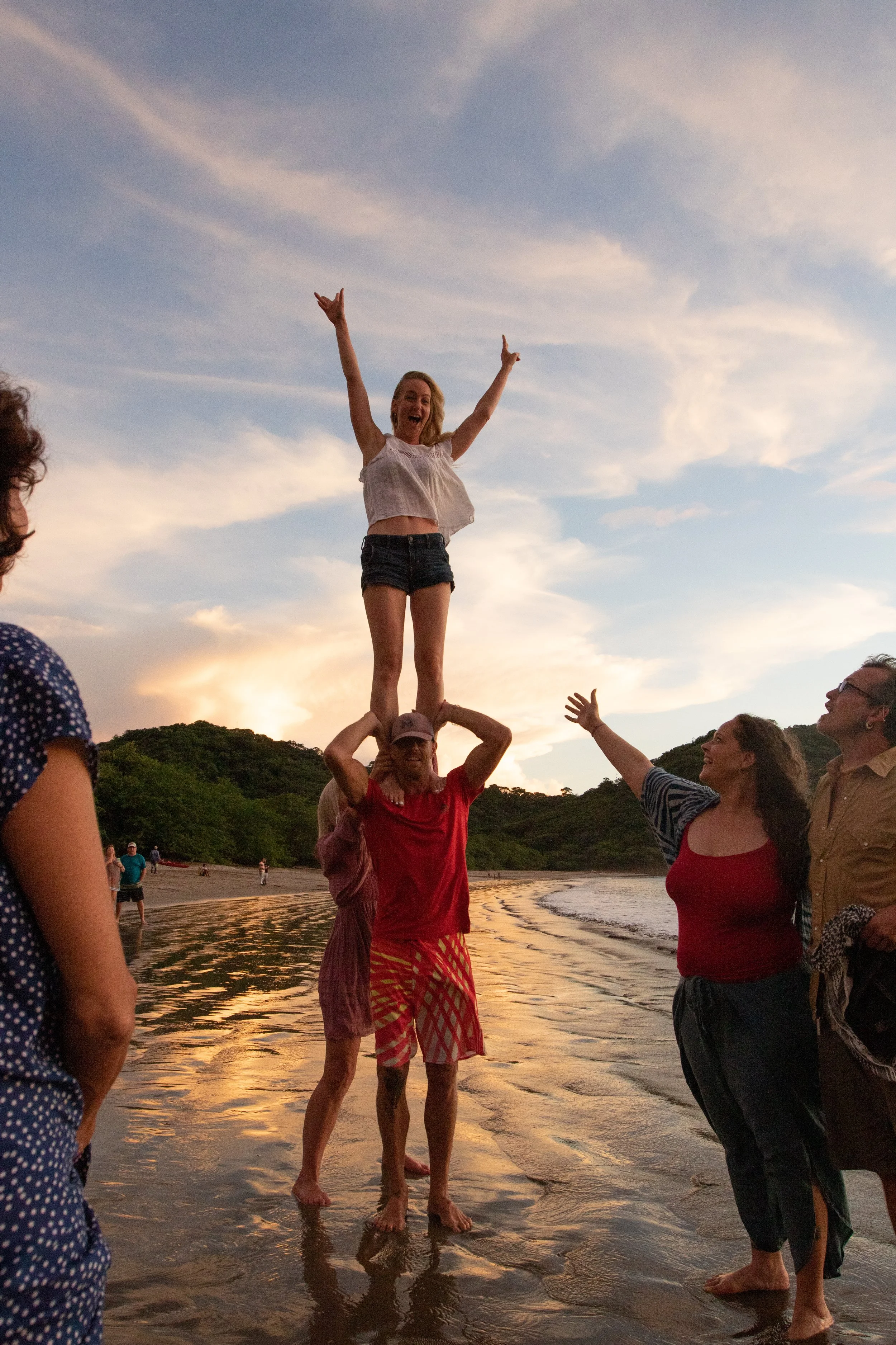 A group of people at the beach during sunset, with a woman standing on a man's shoulders, smiling and raising her arms in a cheer. Others around are watching and enjoying the moment.