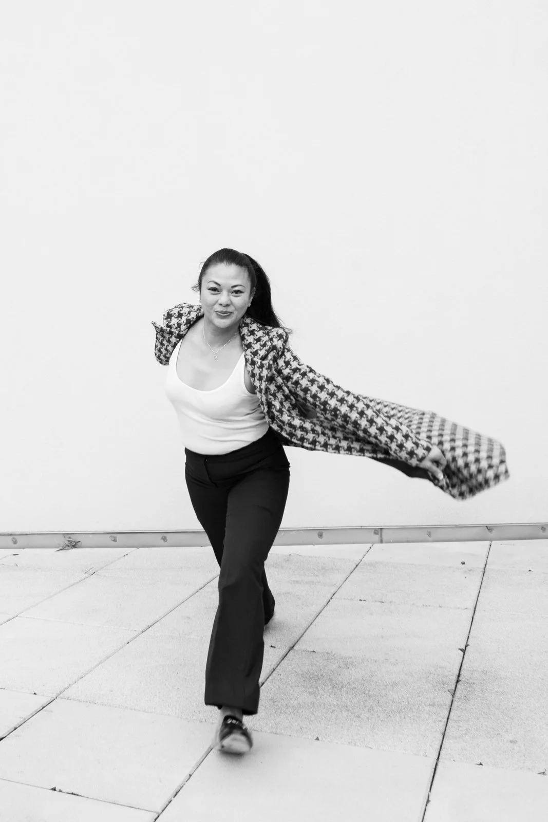 A woman with long hair pulled back, wearing a patterned jacket, white tank top, dark pants, and sneakers, is walking on sidewalk in front of a plain white wall.