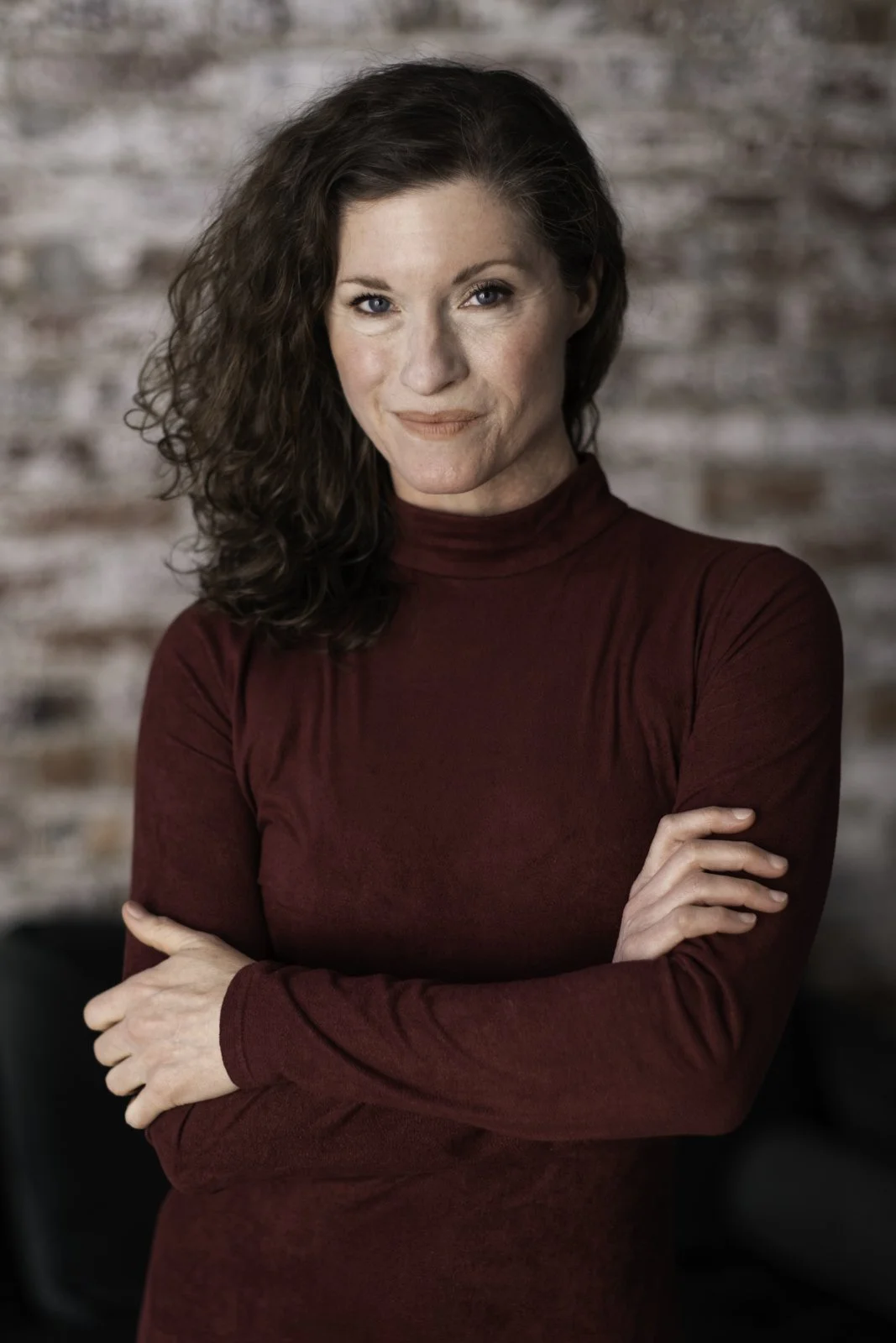 A woman with curly brown hair and blue eyes, wearing a maroon turtleneck, standing with crossed arms in front of a brick wall.