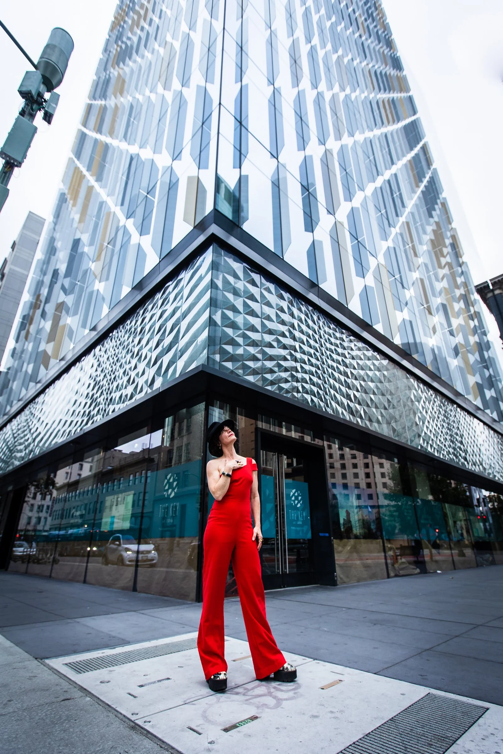 A woman wearing a red jumpsuit and black hat standing outside a modern glass skyscraper in an urban setting.