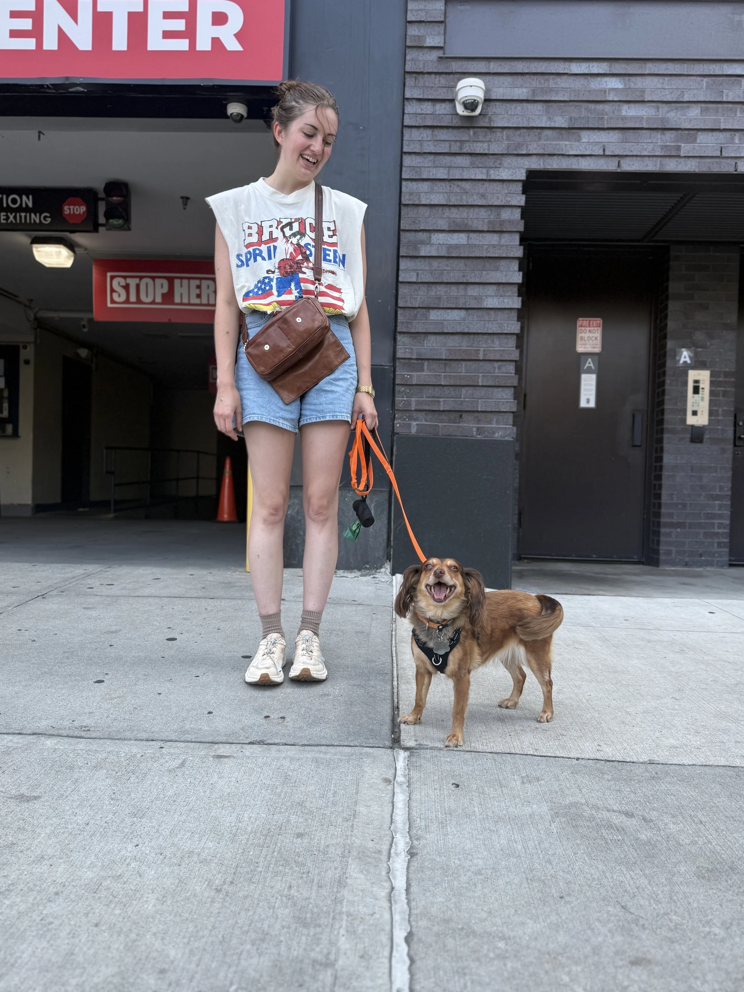 A young woman in a vintage Bruce Springsteen t-shirt and high-waisted denim shorts is standing outdoors on a sidewalk, smiling while holding an orange leash attached to a small brown dog with floppy ears. The dog is wearing a black harness and appears happy. There is a parking garage entrance behind them with red signs that say 'STOP HERE' and 'ENTER', along with a security camera mounted on the wall.