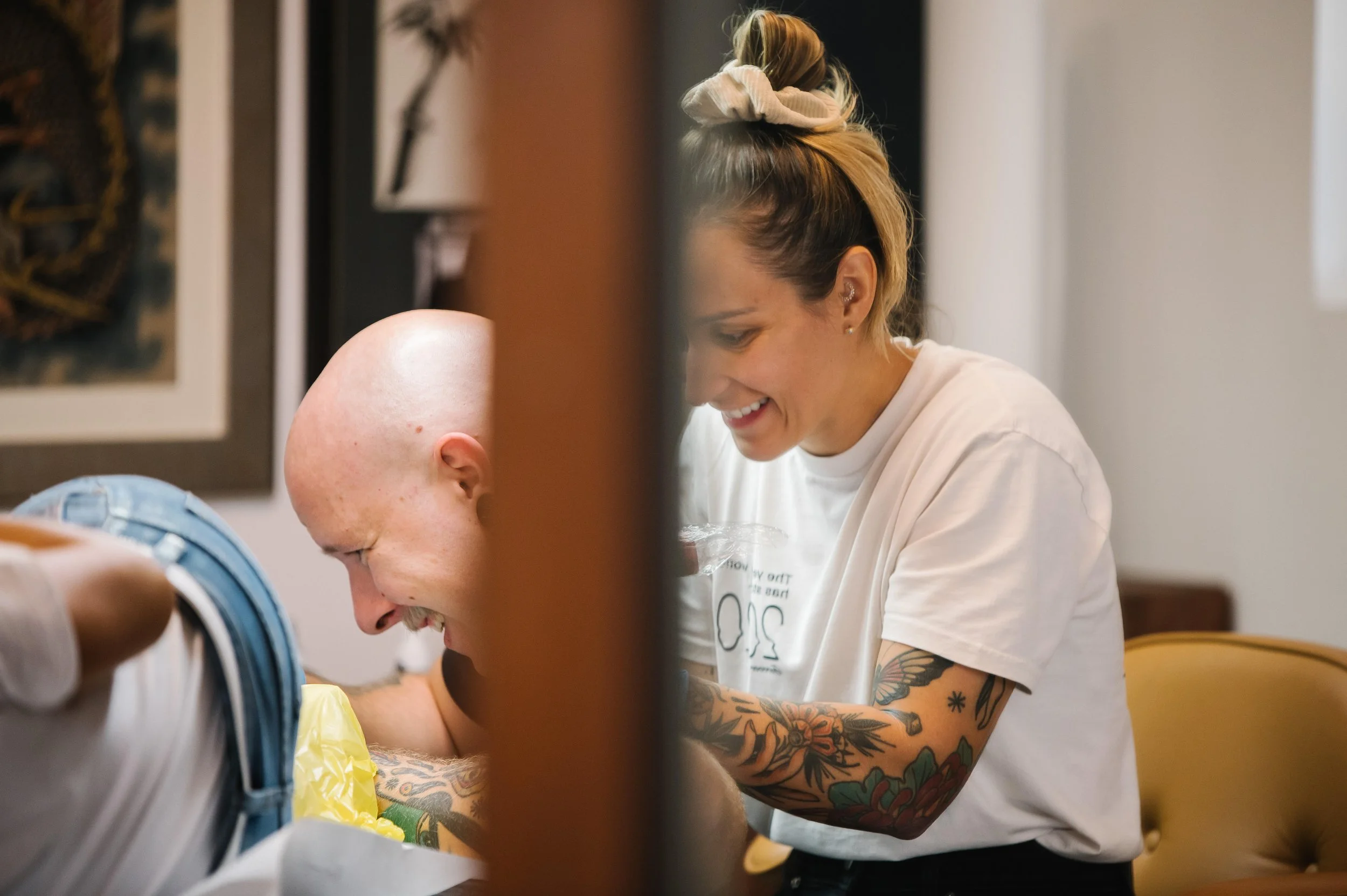 A smiling woman with tattoos on her arms and a bun hairstyle is interacting with a person who appears to be in a medical or caregiving setting. The woman is wearing a white T-shirt with some text, and they are both engaged in a positive moment, possibly sharing a laugh.
