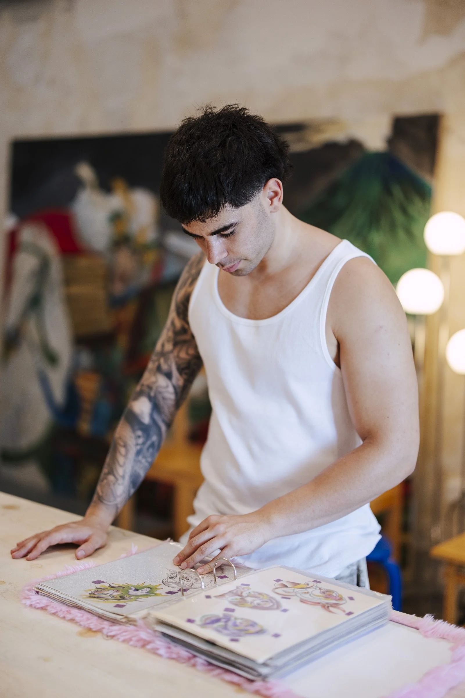 A young man with tattoos on his arm wearing a white tank top is looking down at an open scrapbook or journal on a table, which contains colorful illustrations or stickers. The background shows a blurred interior with artwork and a lamp.