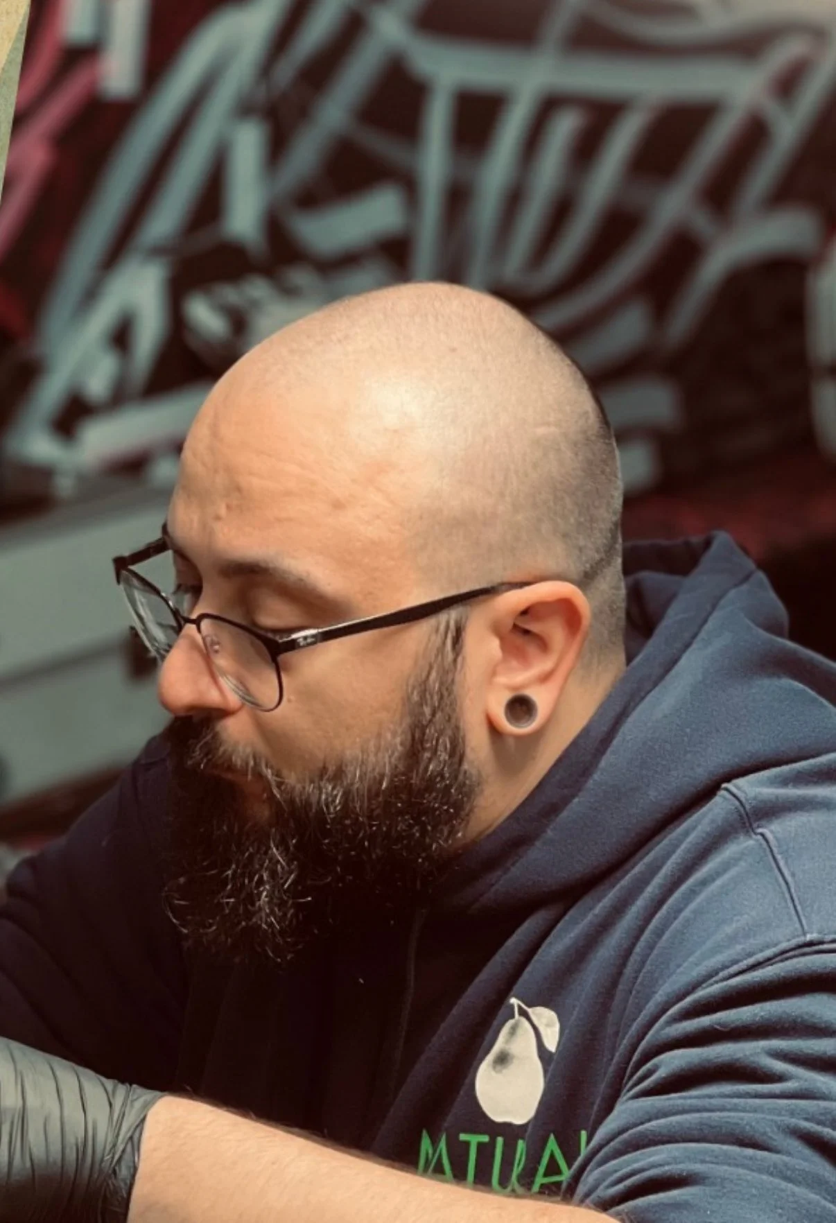 A bald man with glasses, a beard, and ear gauges is sitting and looking down, with a large graffiti-style art piece in the background.