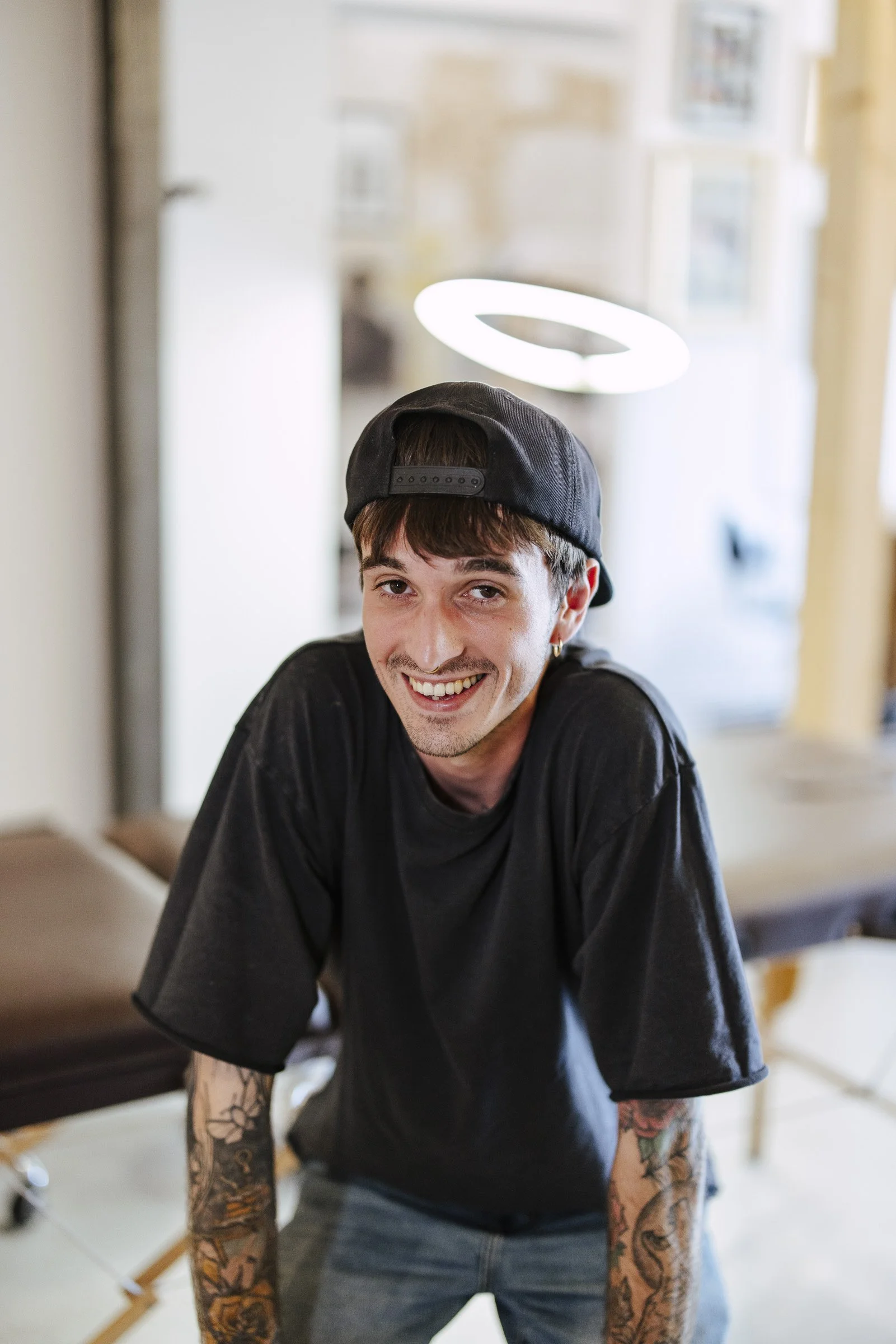 A young man with tattoos on his arms is wearing a black cap backwards and a black T-shirt. He is smiling and leaning forward in a room with a bright background, including a ring light in the distance.