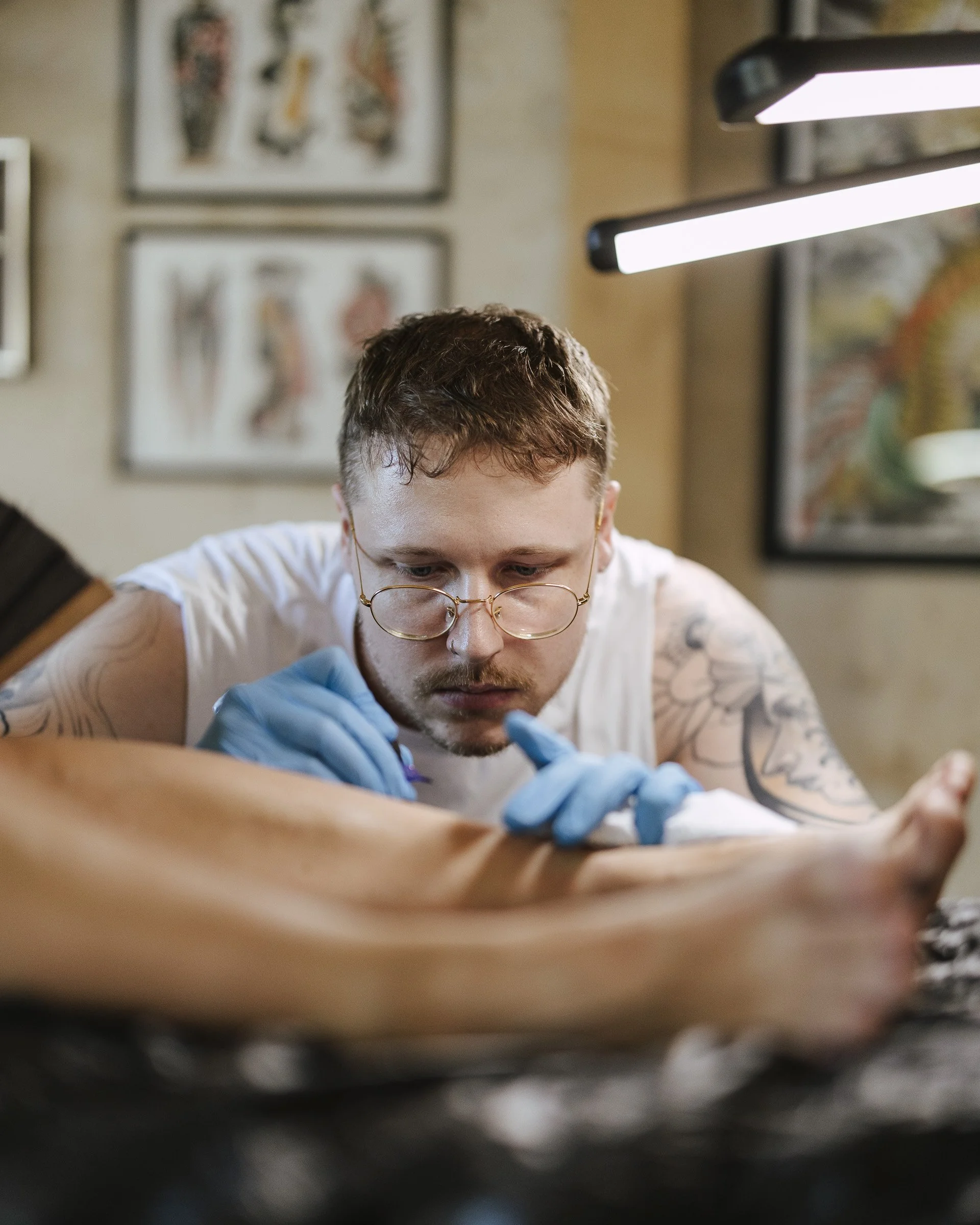 A person with glasses getting a tattoo on their arm at a tattoo studio. The person is looking down and wearing blue gloves. Various framed artworks are visible on the wall behind them.