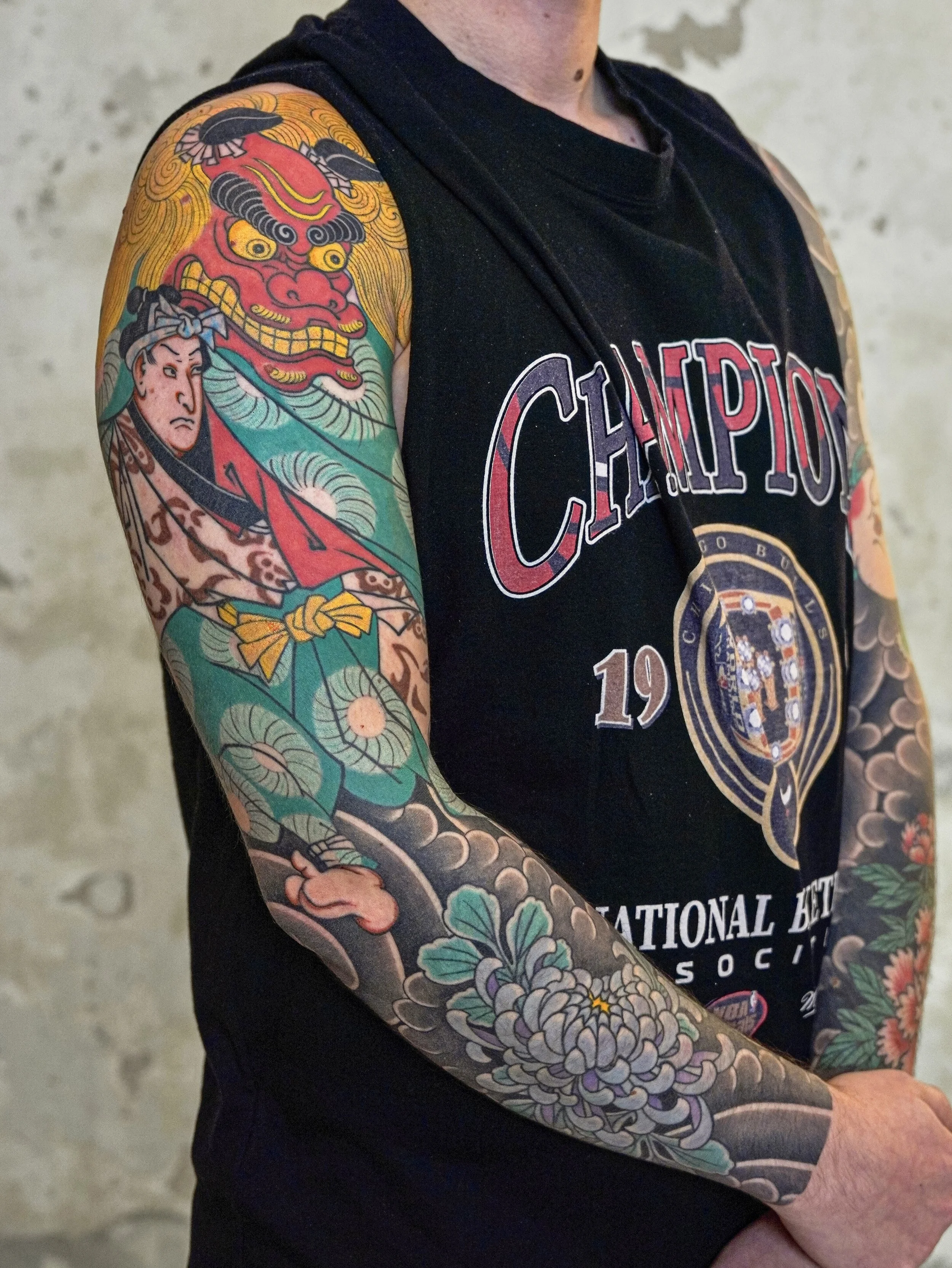 A person with a tribal themed sleeve tattoo on their right arm, wearing a black sleeveless shirt with the word 'CHAMPION' and a logo printed on it, standing against a blurry concrete background.