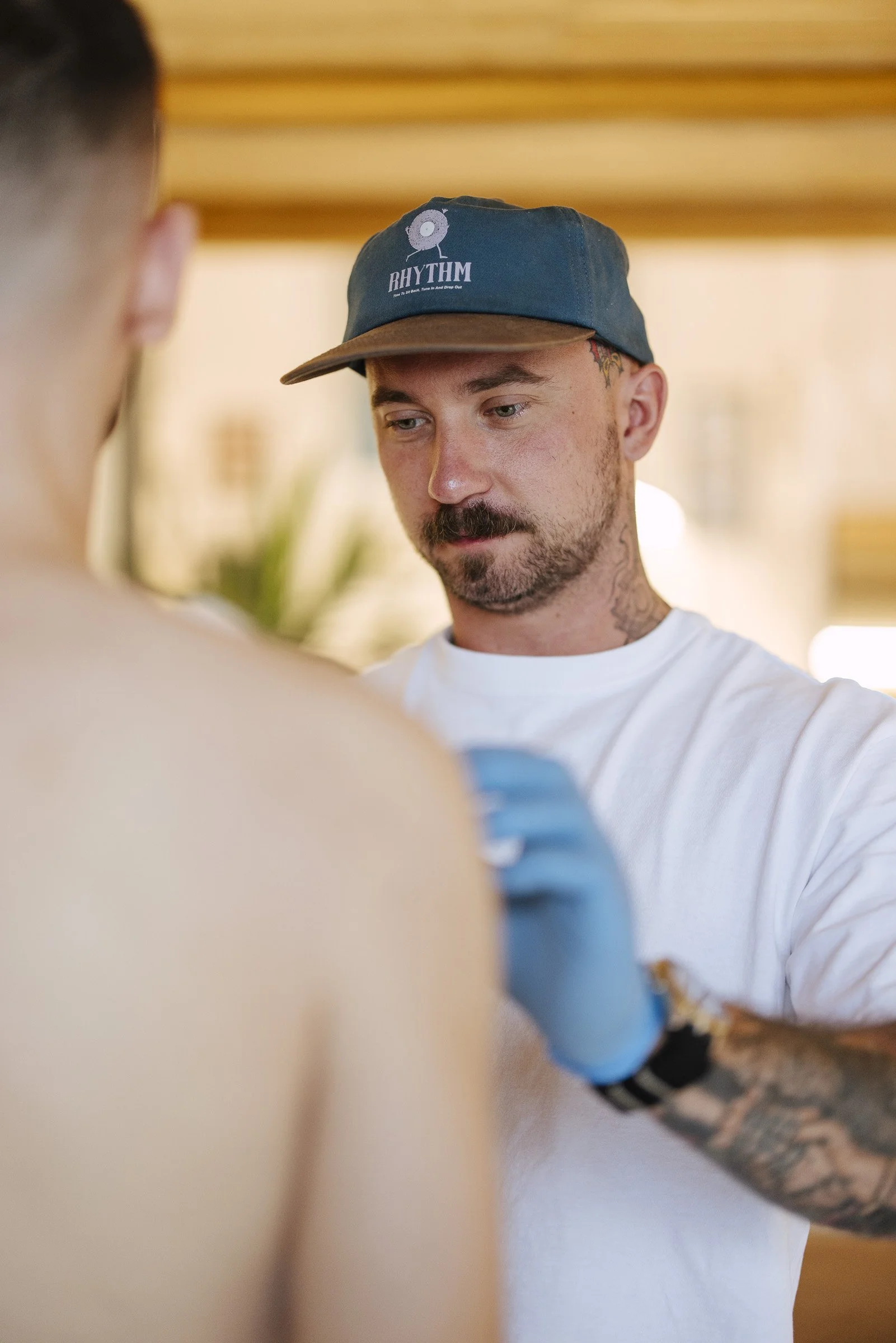 A man wearing a dark baseball cap with the word 'RHYTHM' and a circular logo, and a white t-shirt. He has tattoos on his neck and arm, and a beard. The background is indoors with wooden beams.