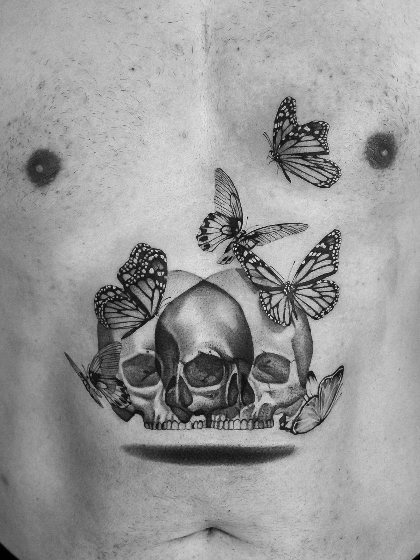Black and white tattoo of a human skull with multiple butterflies flying around and perching on it, located on a person's chest.