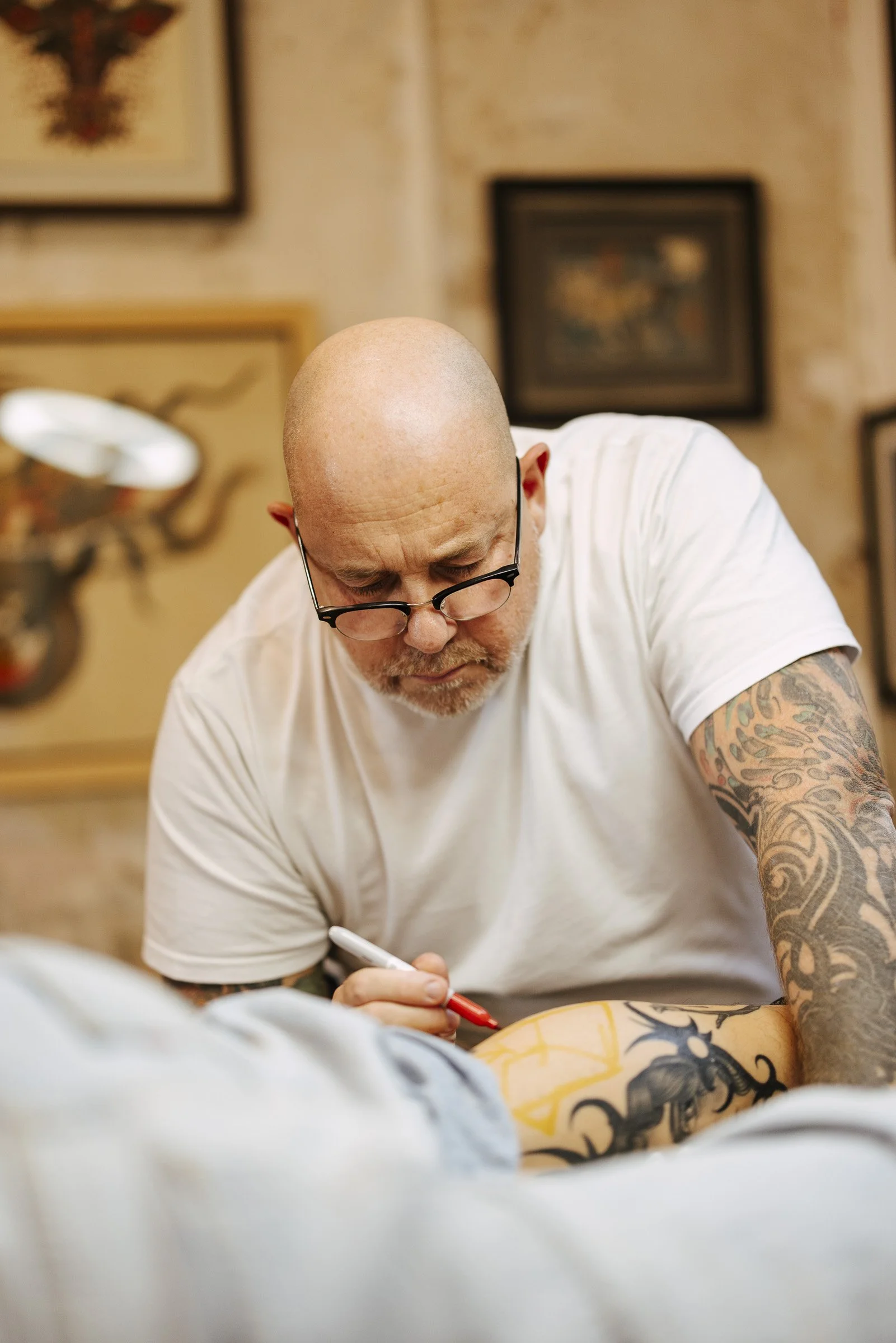 A bald man with glasses and tattoos on his arms is tattooing a tattoo on another person's leg with colorful ink and a marker.