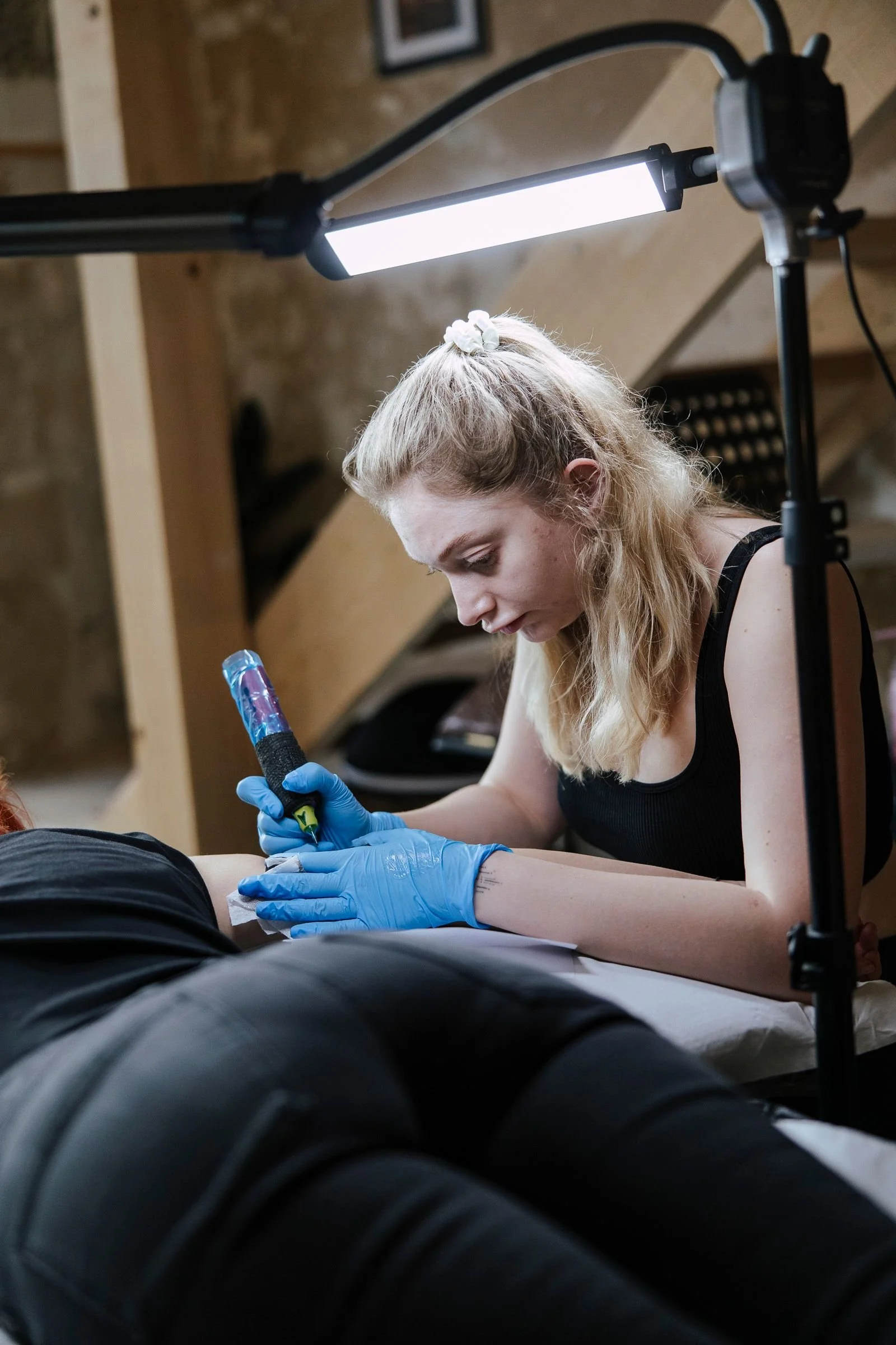 A woman with blonde hair, wearing a black sleeveless top and blue gloves, tattooing a person's arm in a tattoo studio illuminated by a bright overhead light.