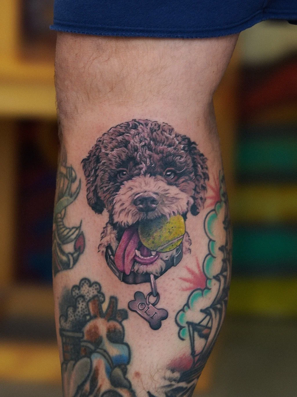 Tattoo of a dog holding a tennis ball in its mouth, with a collar and dog tag, on a person's upper leg.