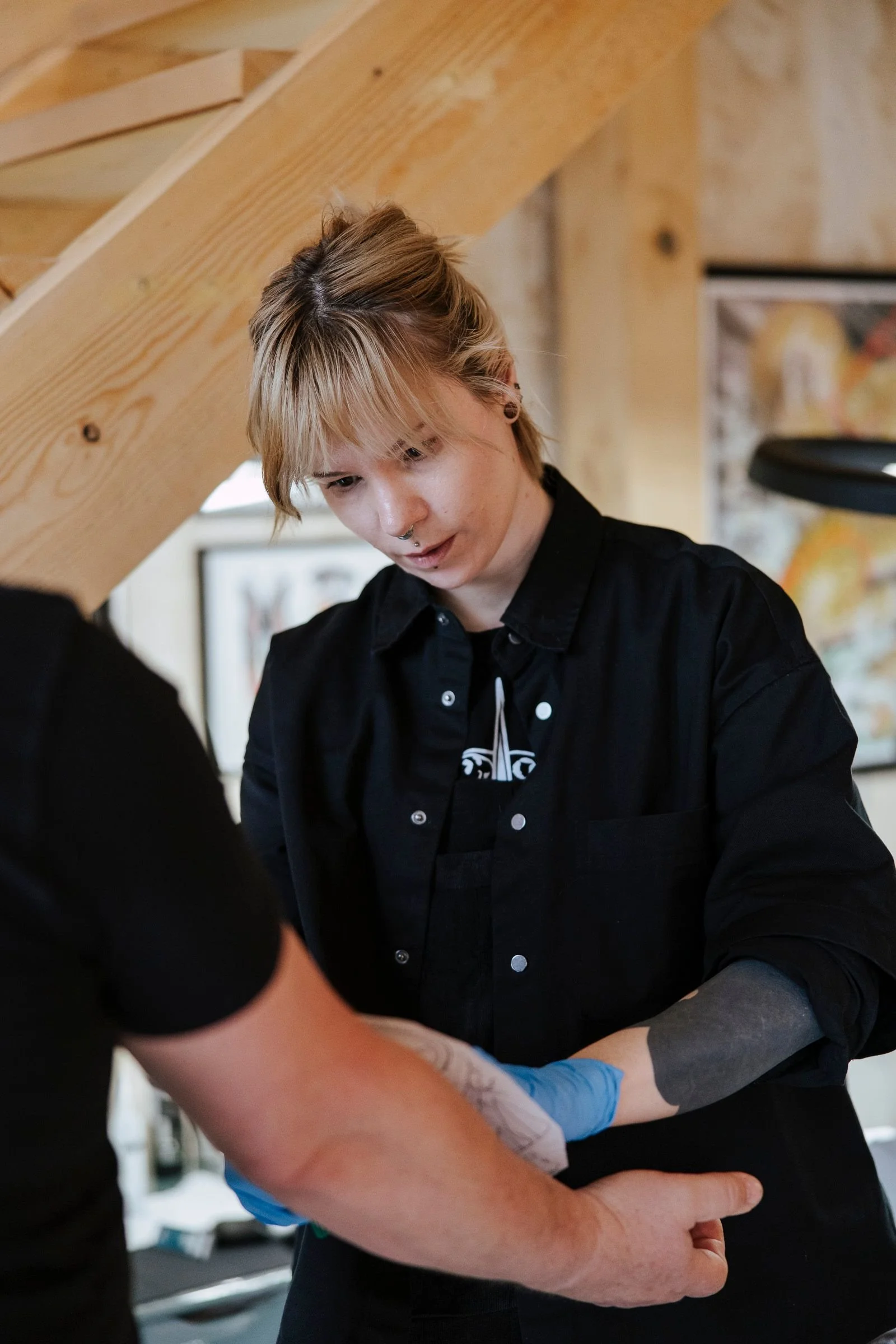 A person with a short blonde haircut and septum piercing, wearing a black shirt, appears to be receiving a tattoo or medical procedure on their arm from a person wearing blue gloves in an indoor setting with wooden beams and framed artwork on the wall.