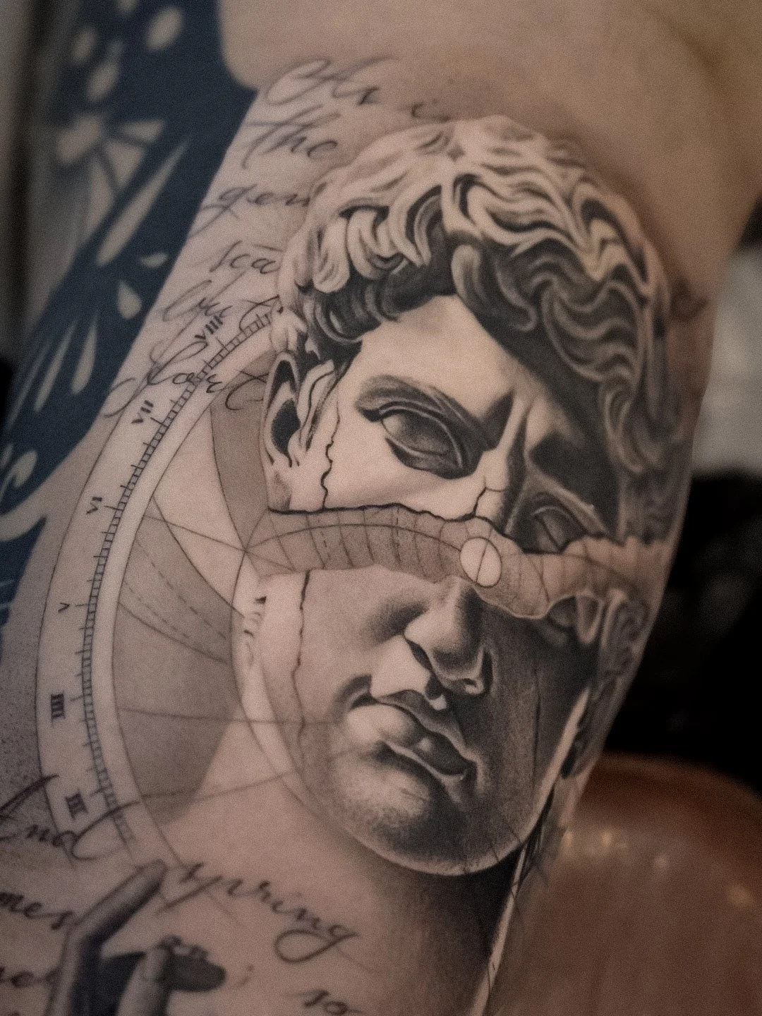 Tattoo of a woman's face with detailed shading and a cracked effect, accompanied by various script writings and geometric elements.