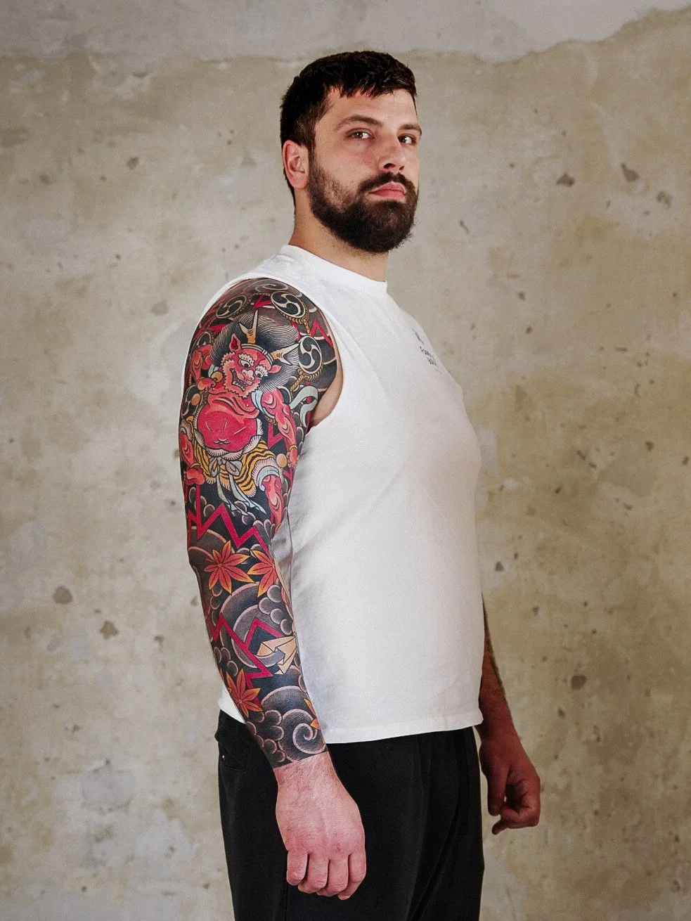 Man with a beard and short dark hair standing sideways against a plain beige wall, wearing a white sleeveless shirt and black pants, displaying a colorful tattoo sleeve on his left arm with traditional Japanese motifs.