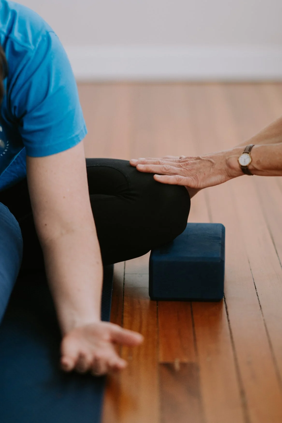 A person practicing yoga, assisted by an instructor, on a yoga mat with their knee touching a block.