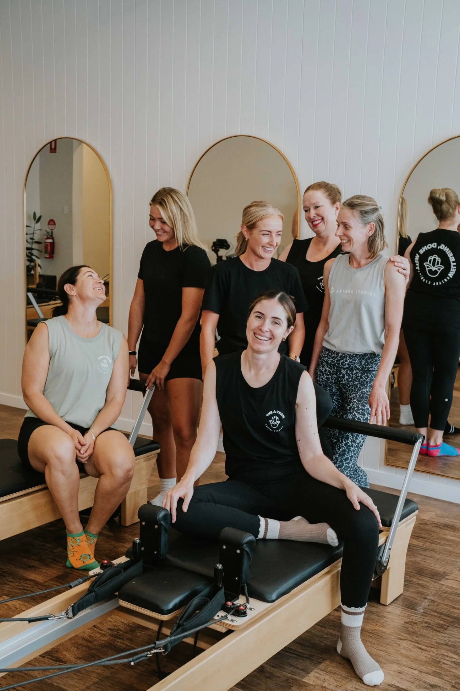 A group of women smiling in front of arched mirrors at Antara Studios, some sitting on Pilates reformer machines and others standing behind them.