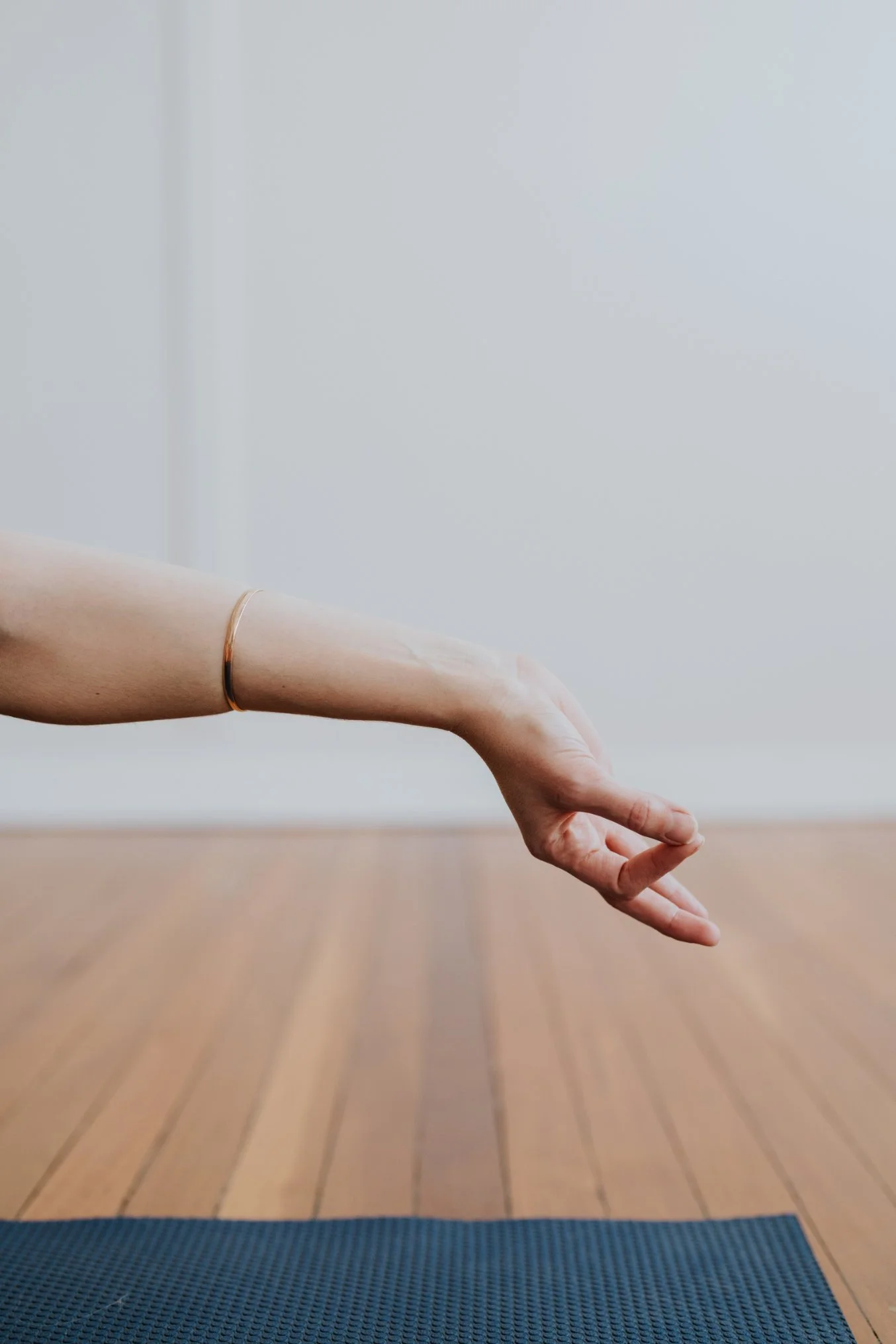 A person's arm reaching forward while doing yoga on a dark yoga mat on a wooden floor. The person is wearing a thin bracelet.