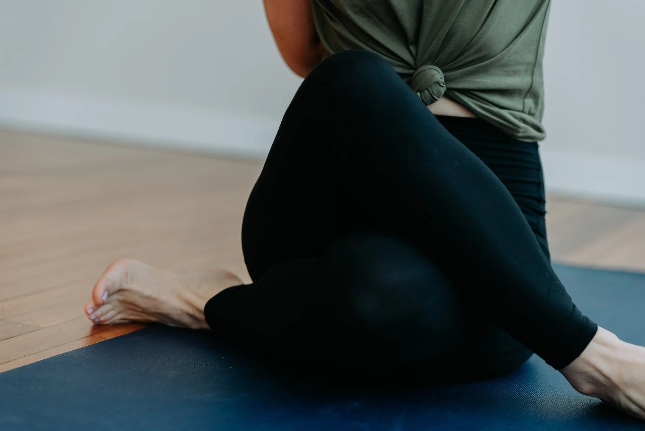 A person practicing yoga or stretching on an exercise mat indoors, wearing black leggings and a green top tied in a knot.