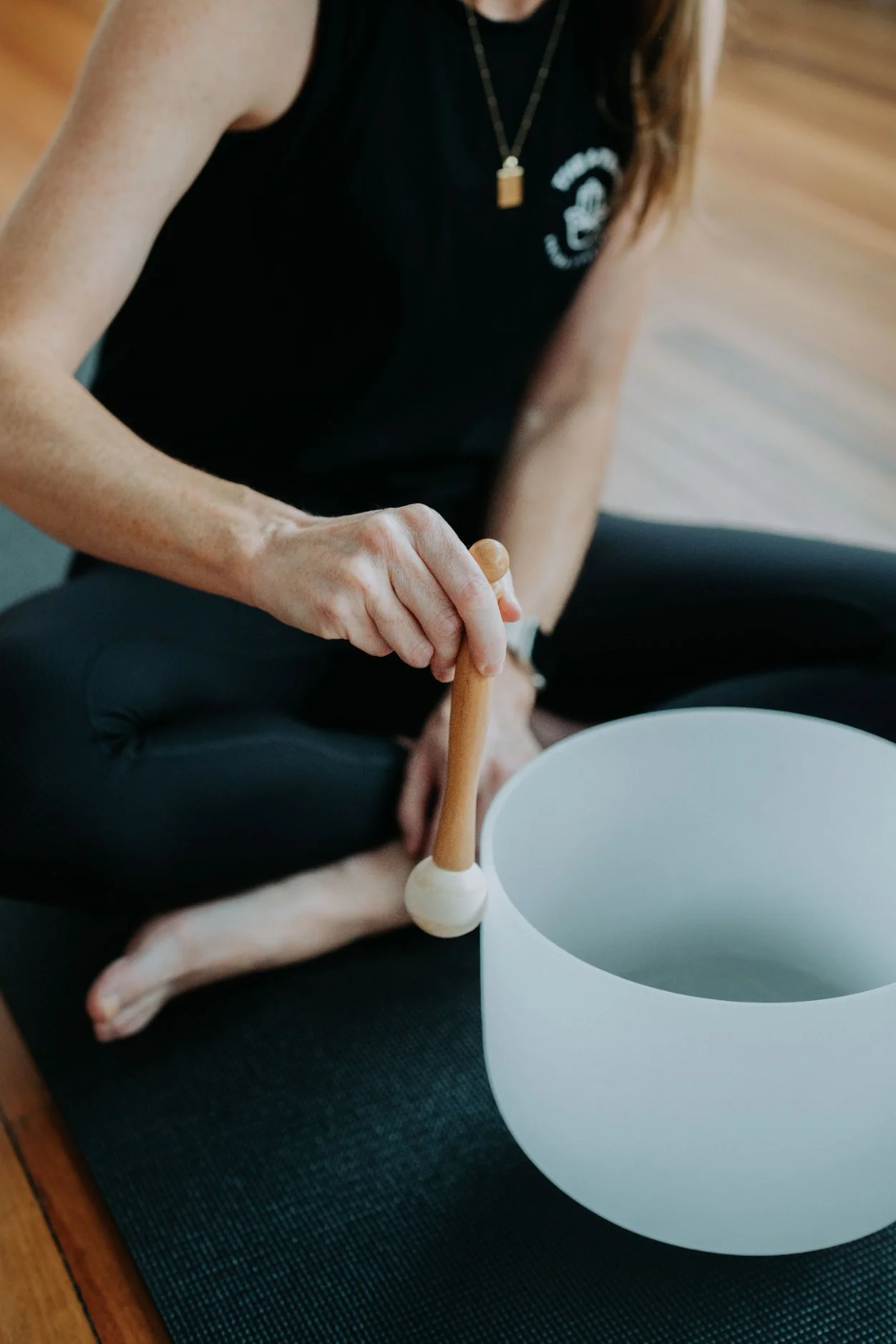 Person sitting cross-legged on a yoga mat, playing a white crystal singing bowl with a wooden instrument.