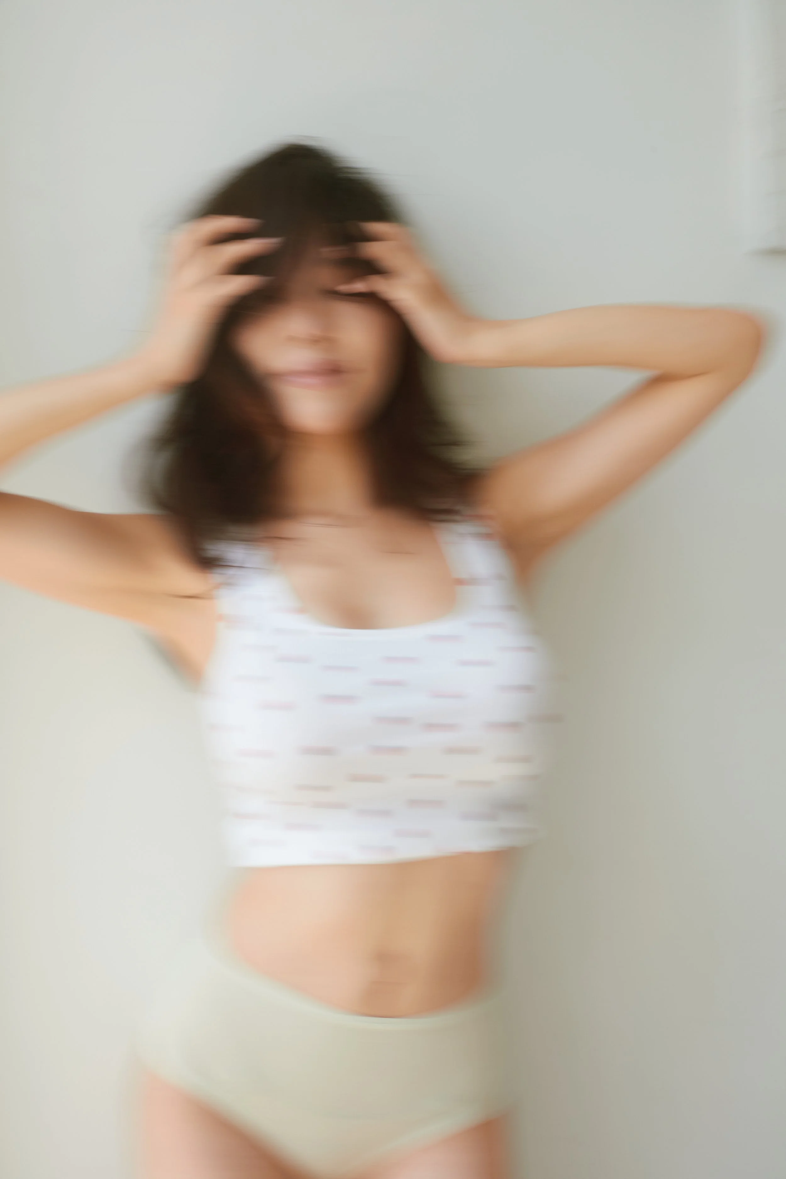 Blurred photo of a woman with dark hair, wearing a white tank top and underwear, holding her head with both hands.
